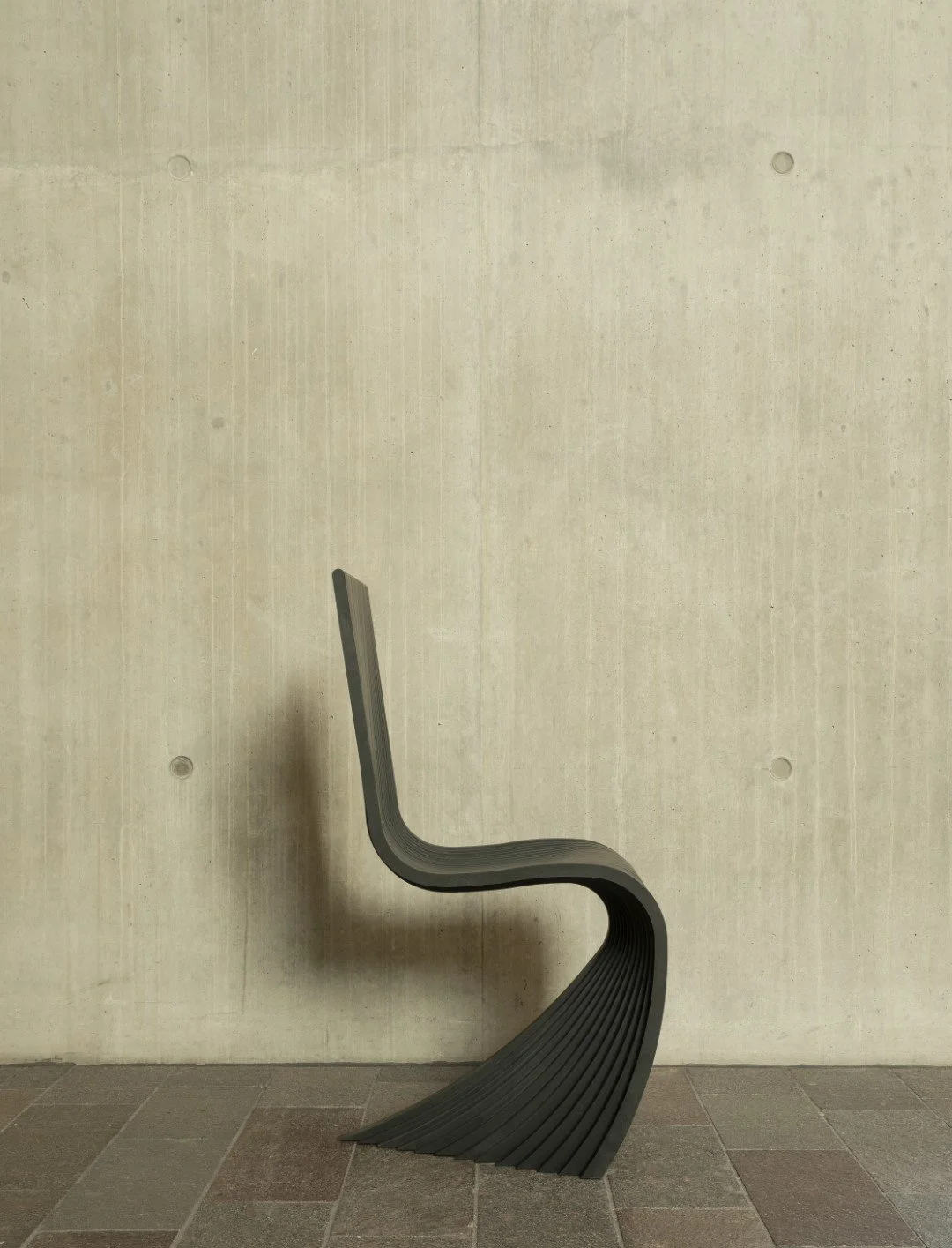 Mike McMahon Studio — Modern black chair with a curved design against a plain beige wall and tiled floor.