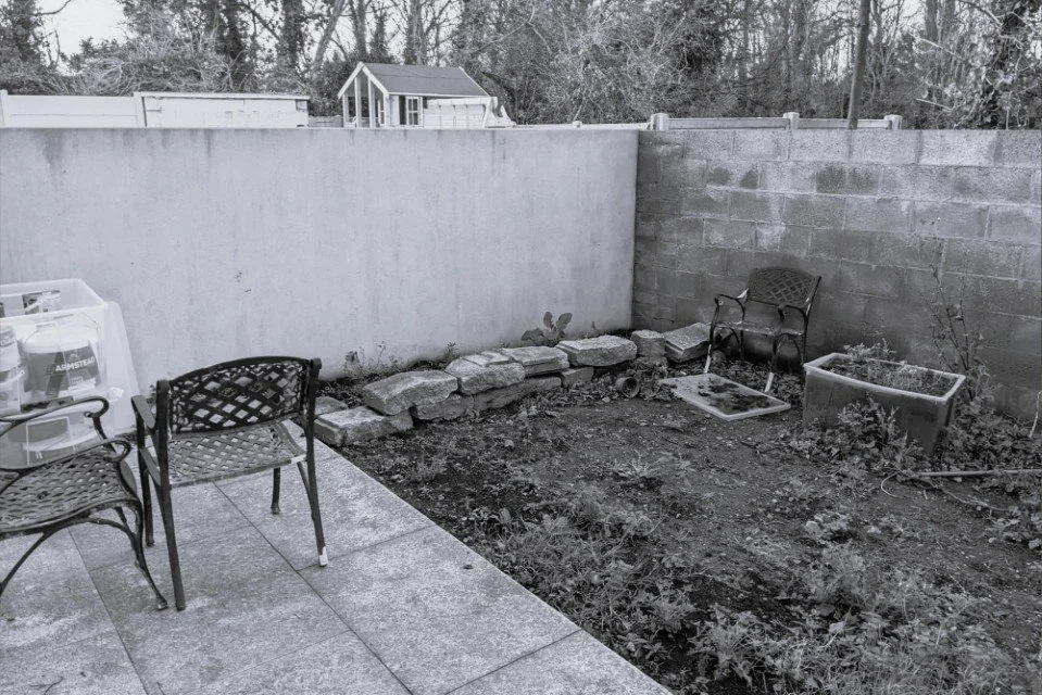 Backyard garden with patio furniture, potted plants, and a small shed in the background, with a tall concrete wall.