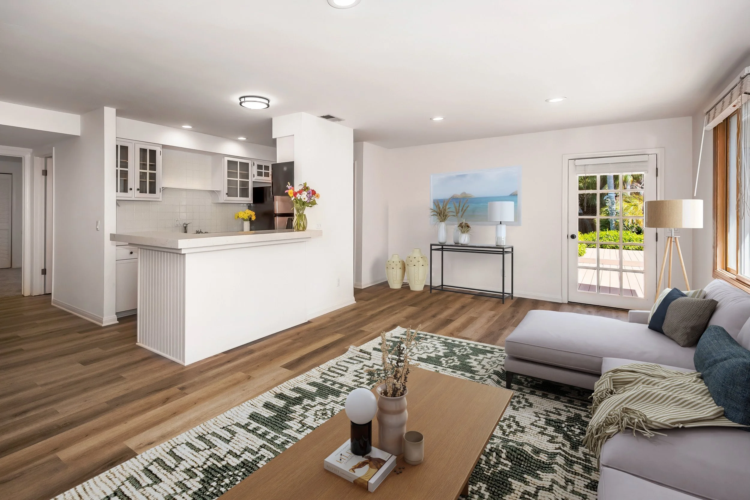 Living room with a white sectional sofa, patterned rug, wooden coffee table with decorative items, and a view of the kitchen with white cabinets and a breakfast bar, open to an outdoor patio. 2426 Shelby B Summerland
