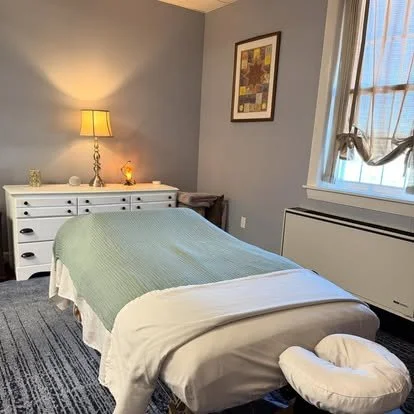 A treatment room with a massage table covered in a light green blanket, a dresser with a lamp, and a picture hanging on the gray wall. There is a window with a curtain and some objects on the dresser.