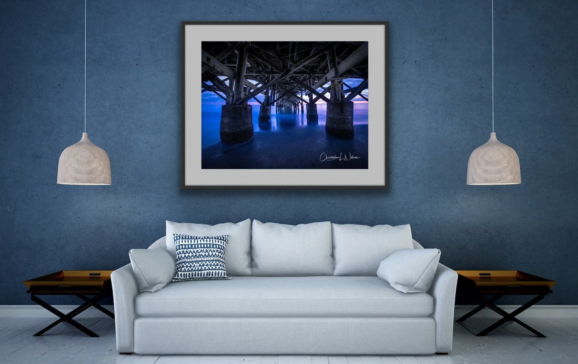 Living room with a white sofa, blue wall, framed artwork of an under-bridge view at sunset, two hanging lamps, and side tables with a wooden top and black metal legs.