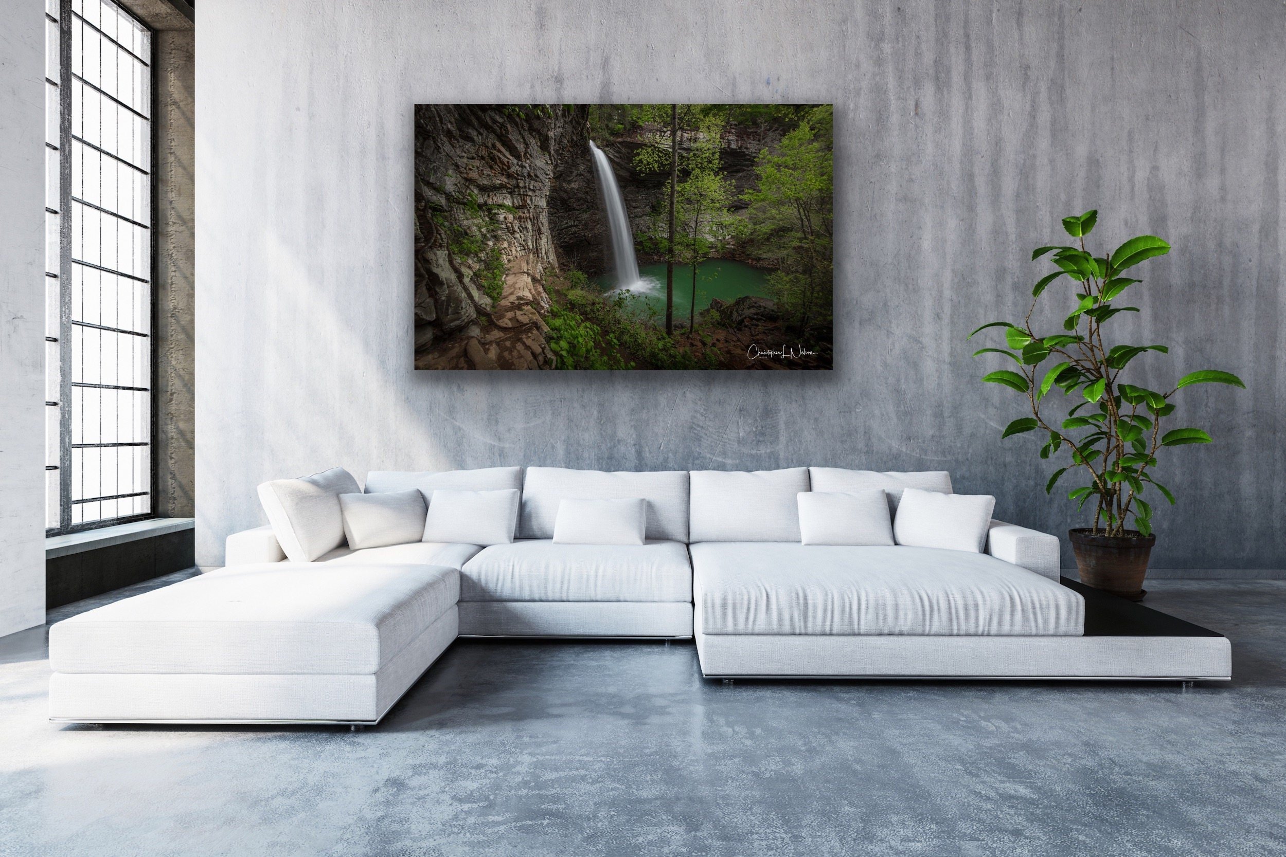 Modern living room with a large white sectional sofa, a potted green plant, and a landscape photograph of a waterfall and forest on the wall.