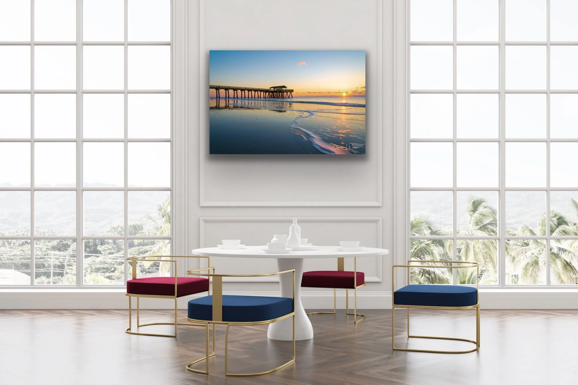 Bright dining room with large windows overlooking palm trees, white walls, a round white table set with white dishes, and four modern chairs with gold frames and red and blue cushions. A landscape painting of a sunset over a pier hangs on the wall.