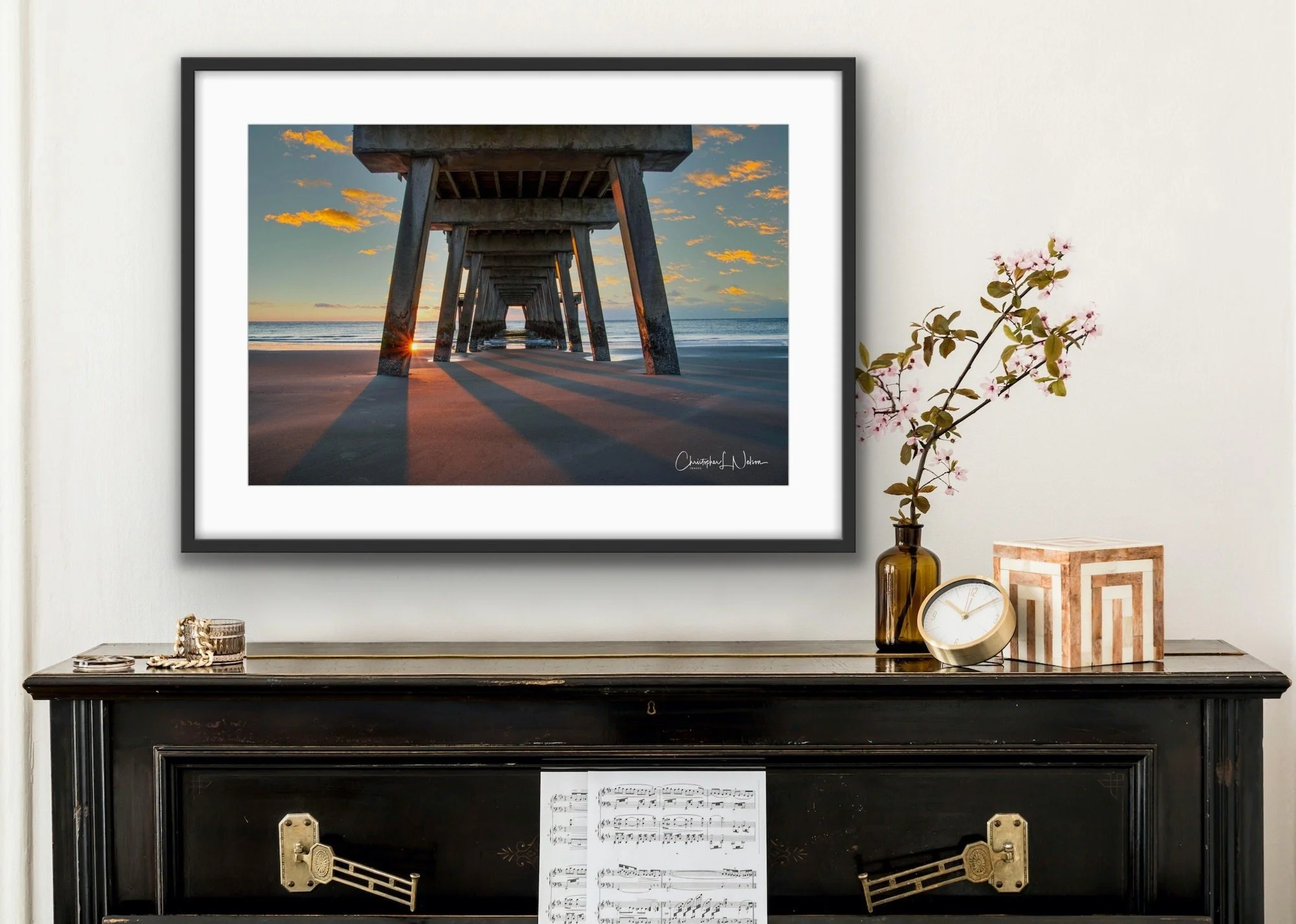 A framed photograph of a pier extending into the ocean during sunset hangs above a black console table decorated with a vase church with pink blossoms, a clock, and a decorative box, placed inside a room.