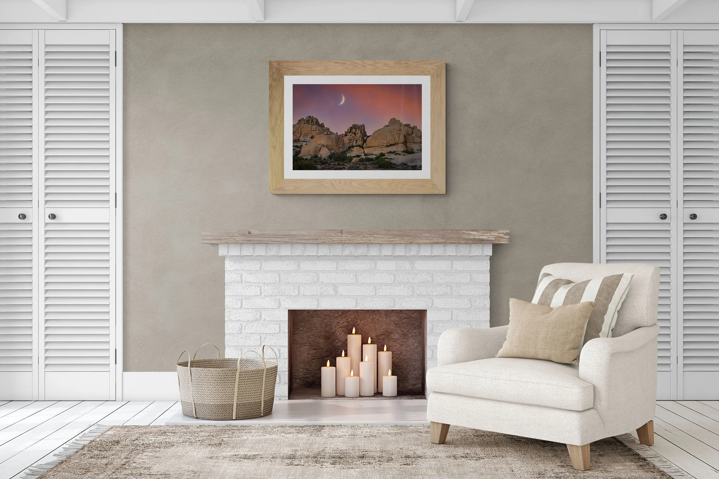 Living room with a white brick fireplace, candles inside, a beige armchair with pillows, a woven basket, a framed landscape photo of a desert with rocks and a sunset, and white closet doors.