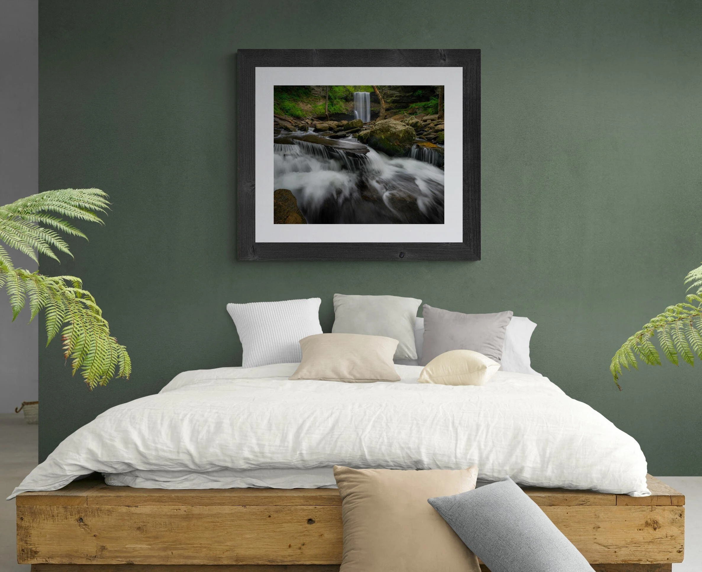 Bedroom with green wall, framed picture of a waterfall, and a wooden bed with white linens and multiple pillows, surrounded by green plants.