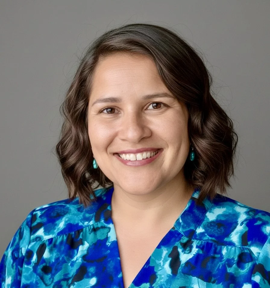 A head shot of Dr. Tati, she is looking at the camera and smiling. She is wearing her hair half up, with coral earrings, a black blazer and multicolor blouse with some coral specks.