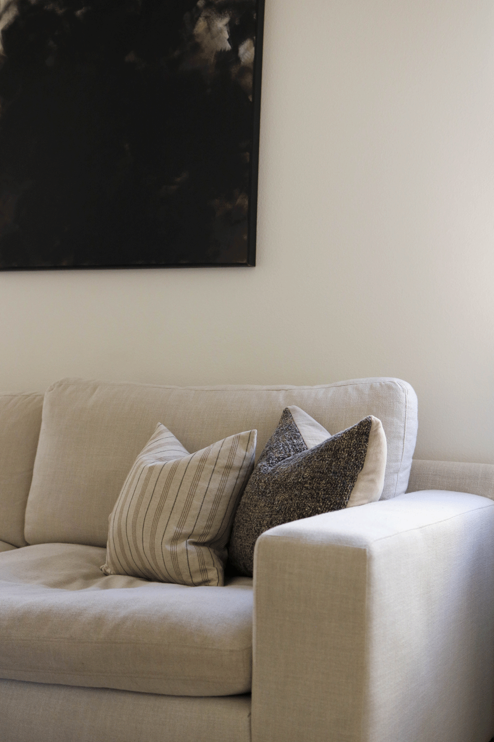A beige sofa with three cushions and a black abstract painting on the wall behind it.