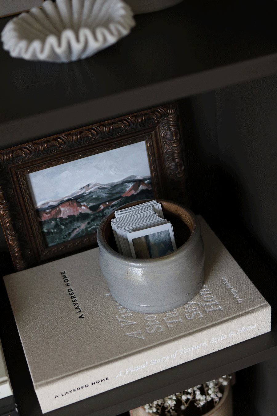 A corner of a shelf with a small framed landscape painting, a stack of postcards or photographs inside a round ceramic bowl, a large beige coffee table book titled 'A Layered Home,' and a white decorative item on top of the shelf.