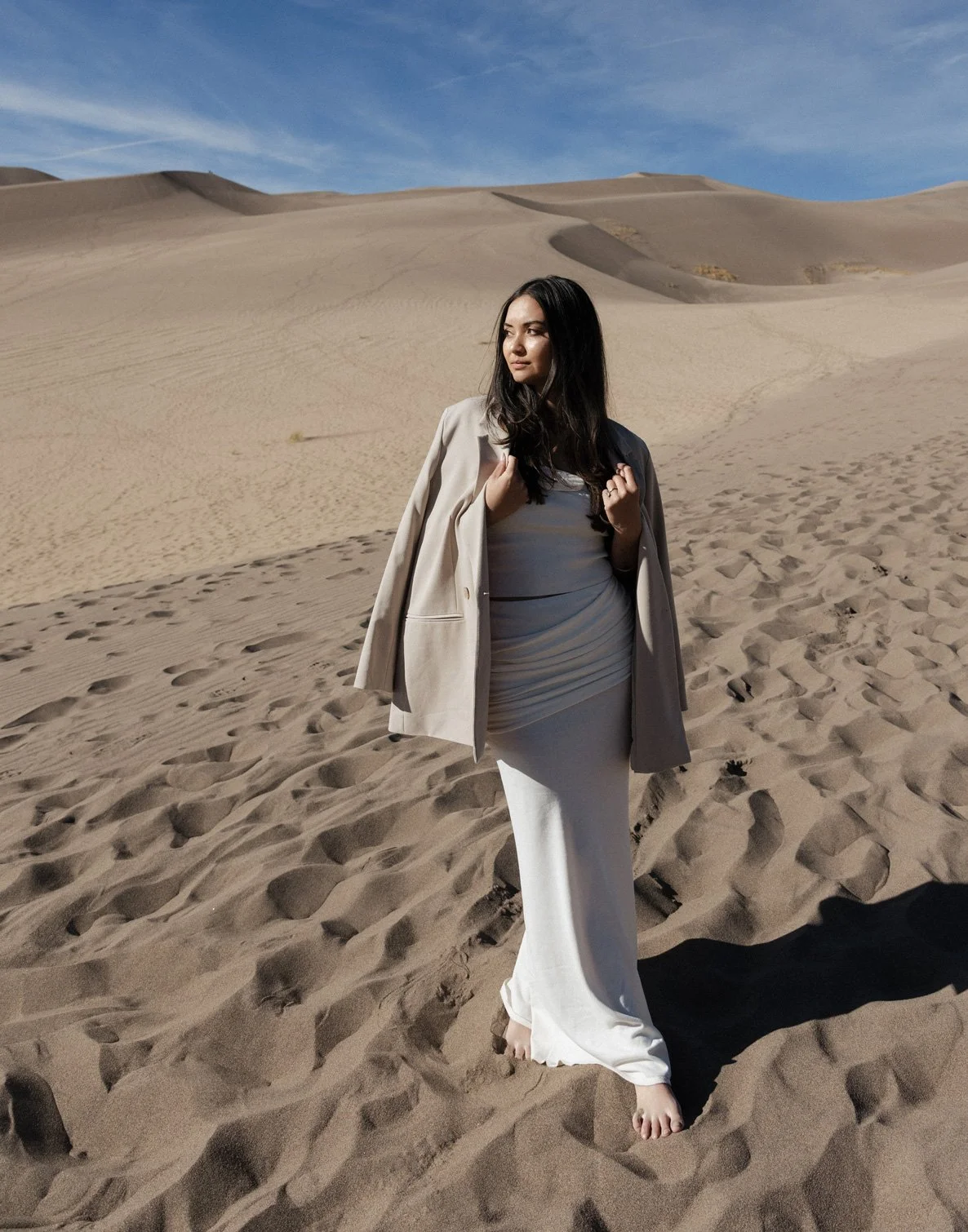 A woman standing barefoot in a sandy desert with dunes in the background, wearing a white dress and a light-colored blazer draped over her shoulders, under a blue sky with wispy clouds.