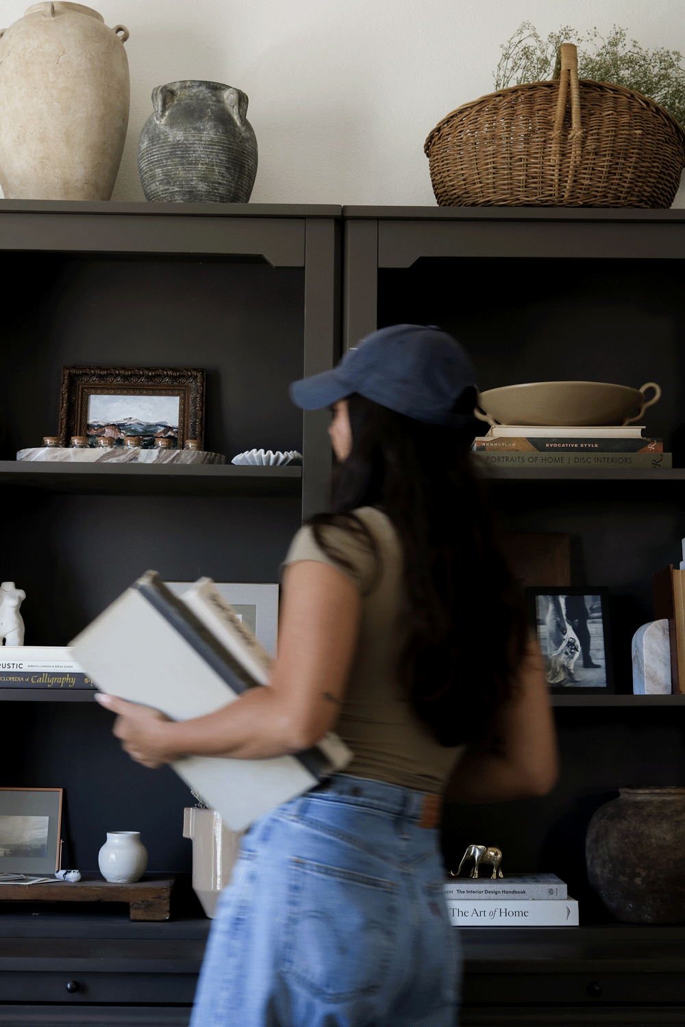 A woman with long dark hair, wearing a blue cap, beige top, and light blue jeans, stands in a room with dark gray shelves holding decorative items, books, and framed photographs, while she holds a stack of books.