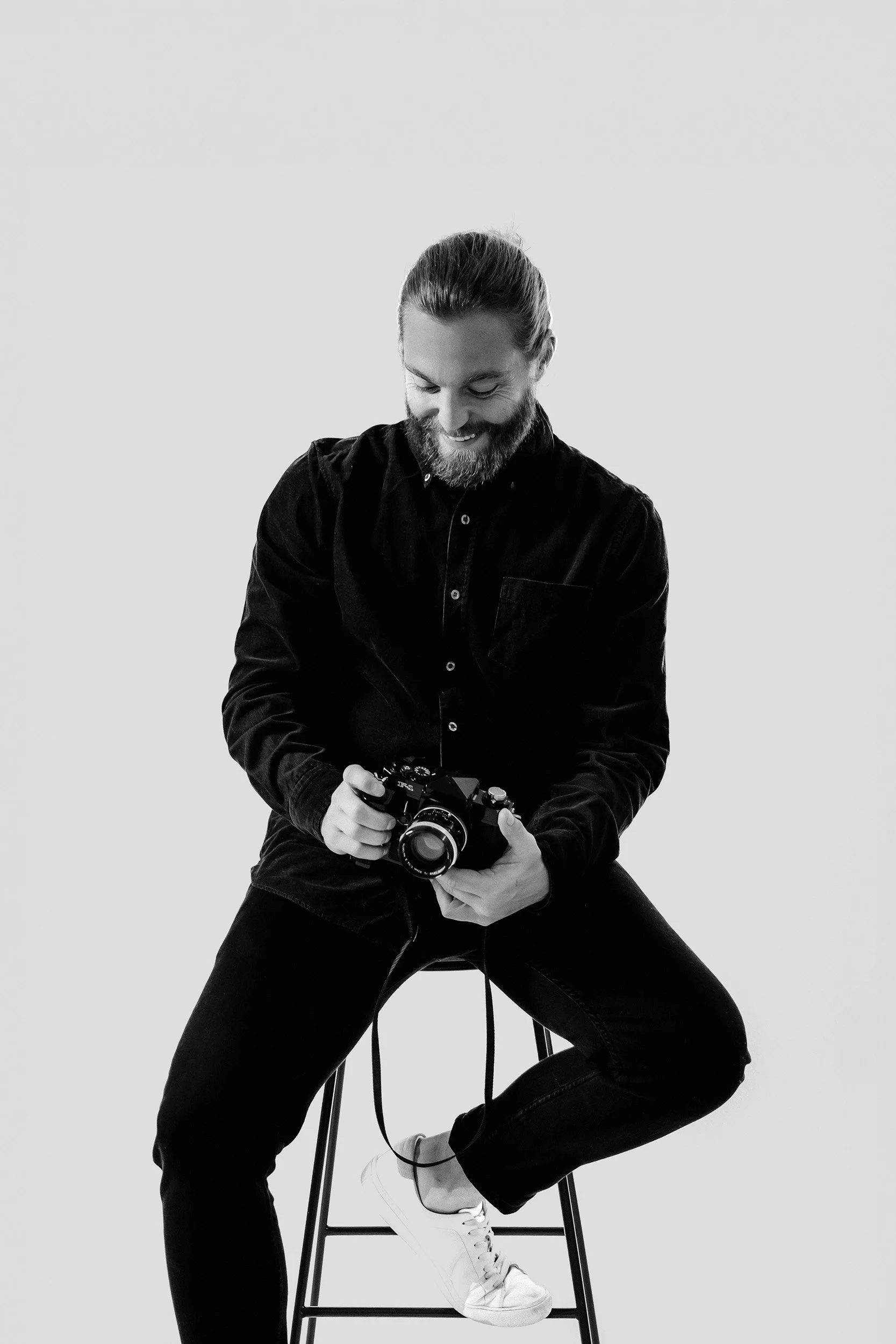 A man with a beard and long hair sitting on a stool, holding a camera, smiling while looking down.