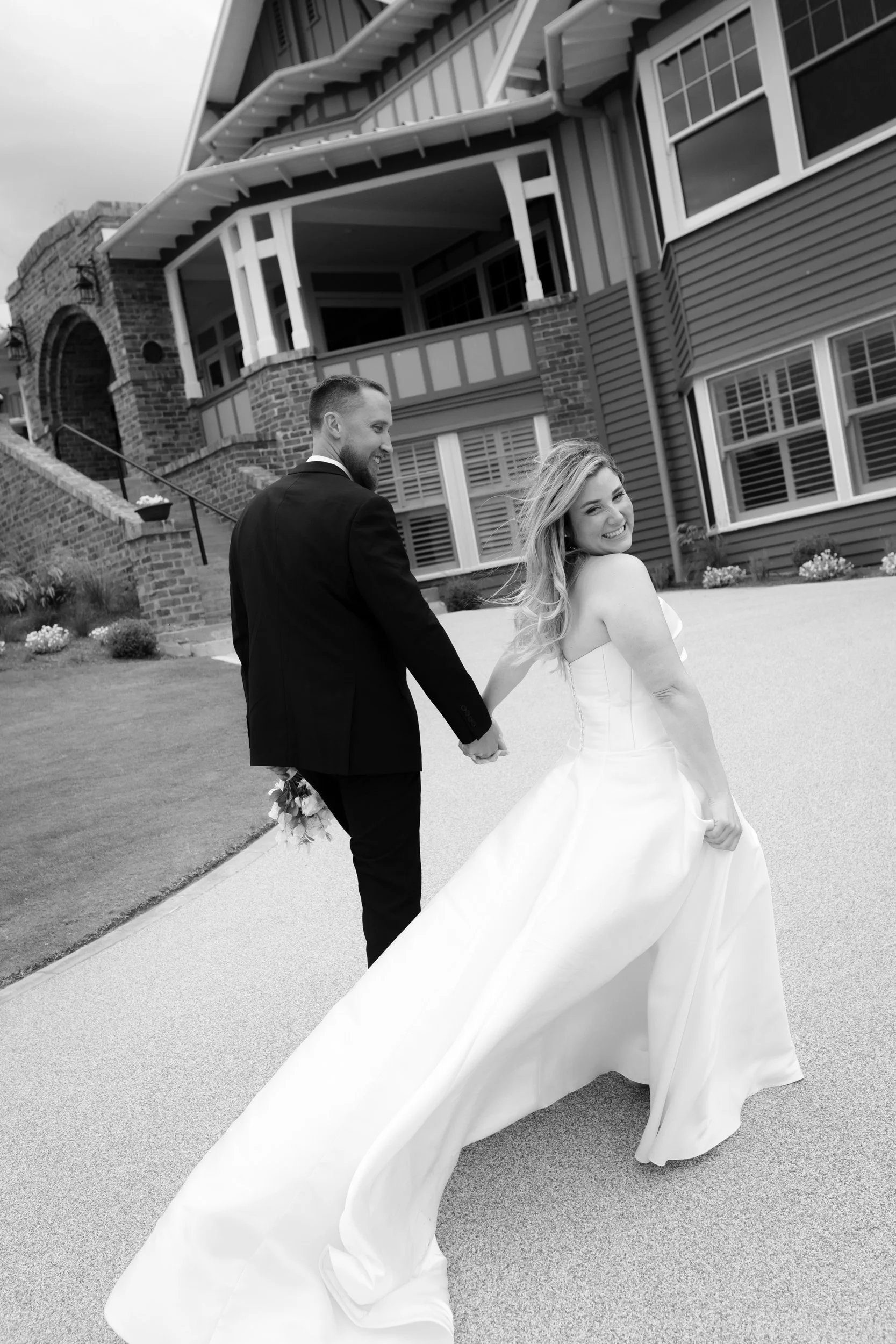Black and white photo of a smiling bride in a strapless wedding gown holding the train of her dress, standing outside the Barwon Heads Golf Course with her husband holding her hand.