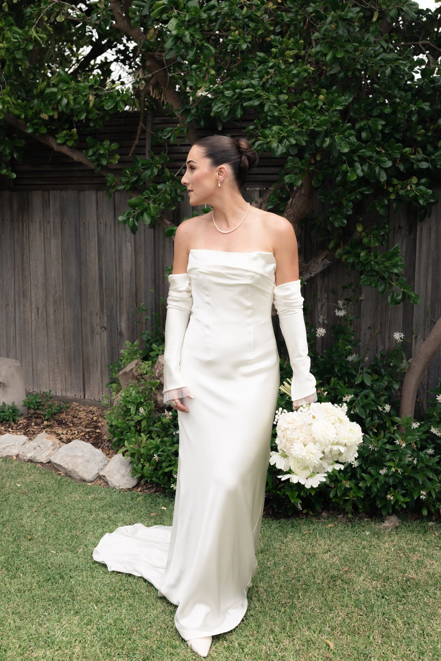 A bride in a white strapless wedding dress with long sleeves and a small train, standing outdoors on grass in front of a wooden fence and green shrubbery, holding a bouquet of white flowers.