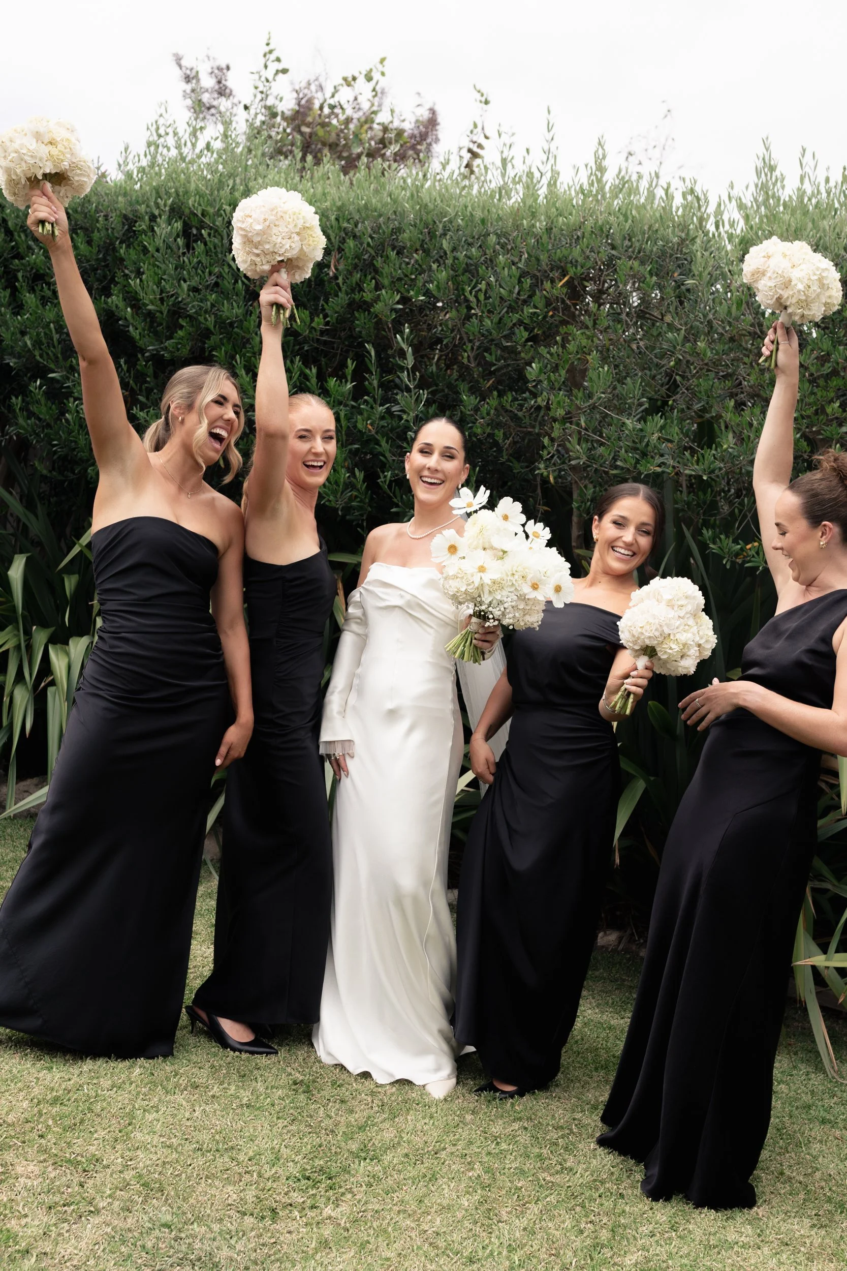A bride in a white wedding dress surrounded by five bridesmaids in black dresses, holding bouquets of white flowers, celebrating outdoors.