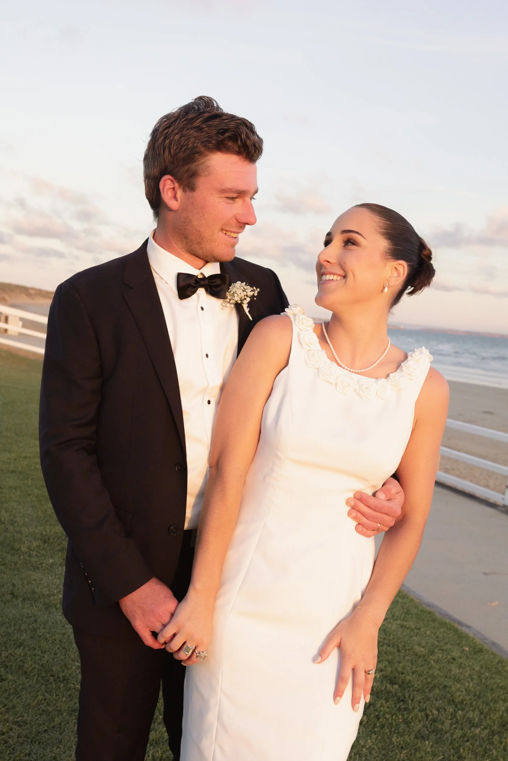 A bride and groom standing close on a beach at sunset, smiling at each other. The groom is wearing a tuxedo with a bow tie and boutonniere, and the bride is in a white dress with a pearl necklace and earrings.