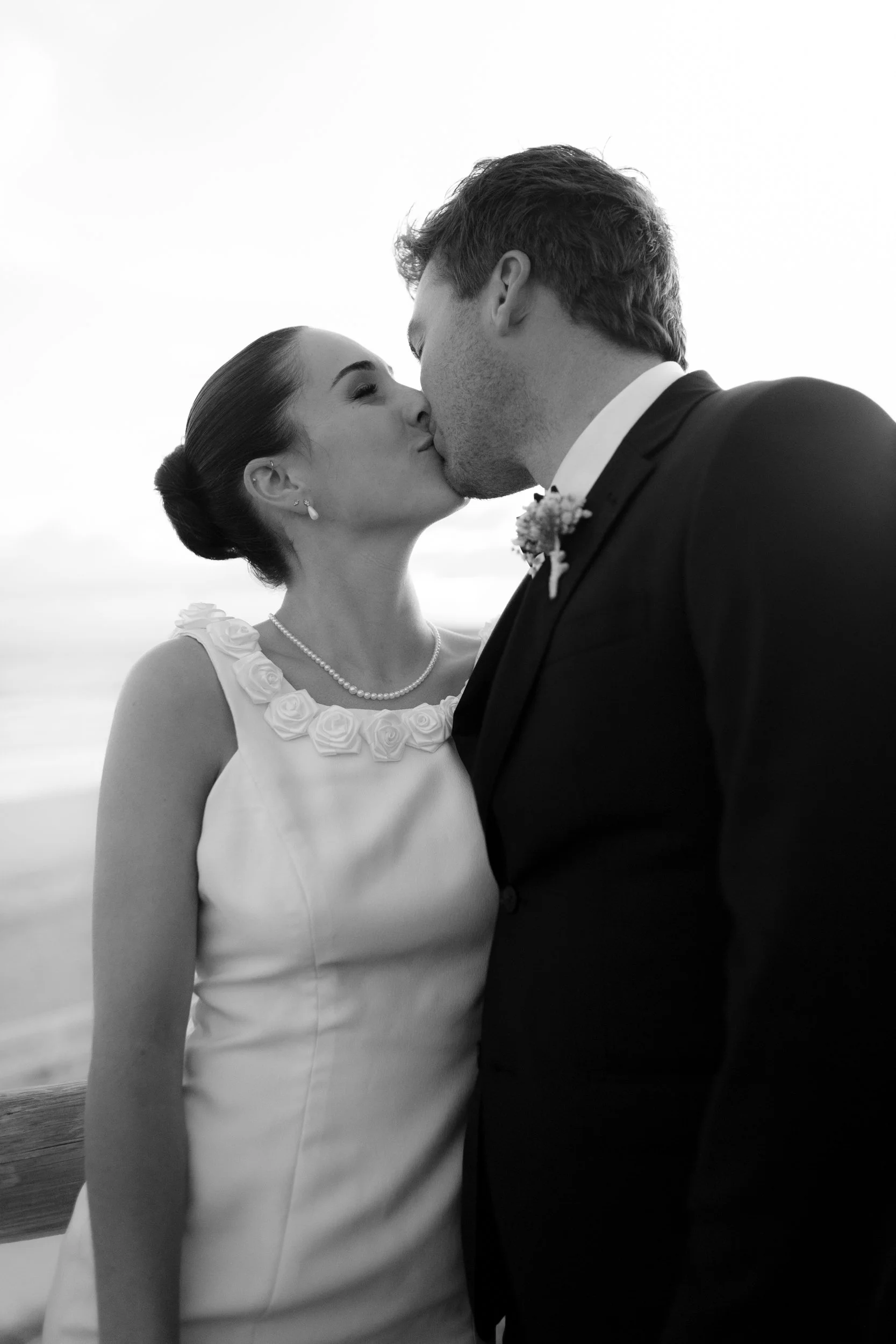 A black and white photo of a bride and groom sharing a kiss on their wedding day. The bride wears a sleeveless dress with flower details around the collar and a pearl necklace, while the groom is in a suit with a boutonniere.