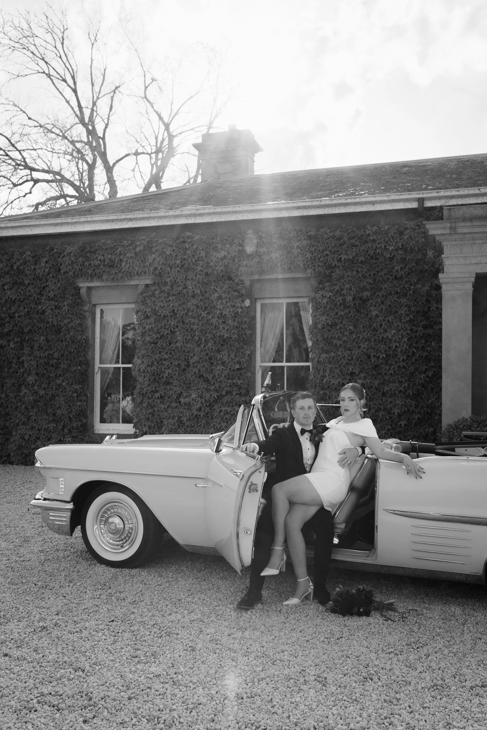 Black and white photo of a man in a tuxedo and a woman in a white dress sitting in a vintage convertible car in front of a large house with ivy-covered walls and tall trees in the background.