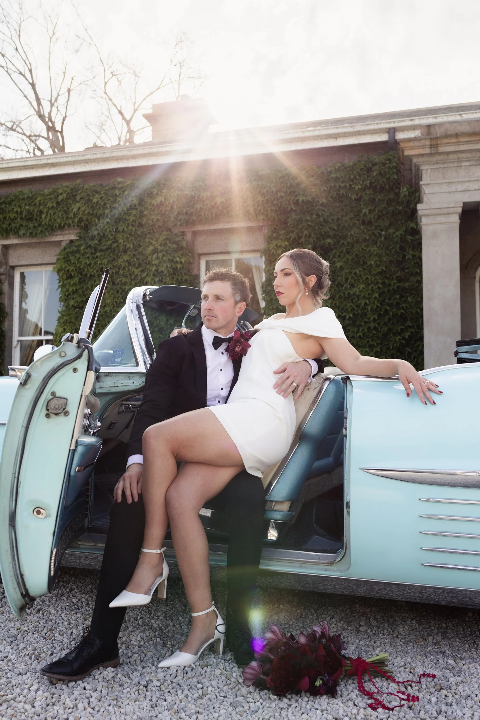 A man in a tuxedo and a woman in a white dress sitting in a vintage car, with the woman resting her arm on the car door, sun shining behind them, and a bouquet of flowers on the ground outside the car.