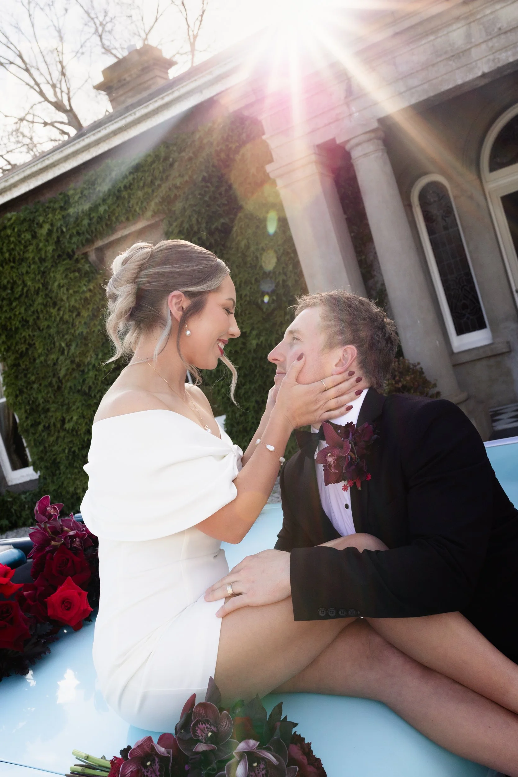 A couple is in a romantic moment during a wedding, sitting in a vintage car with flowers nearby, sun shining brightly from above.