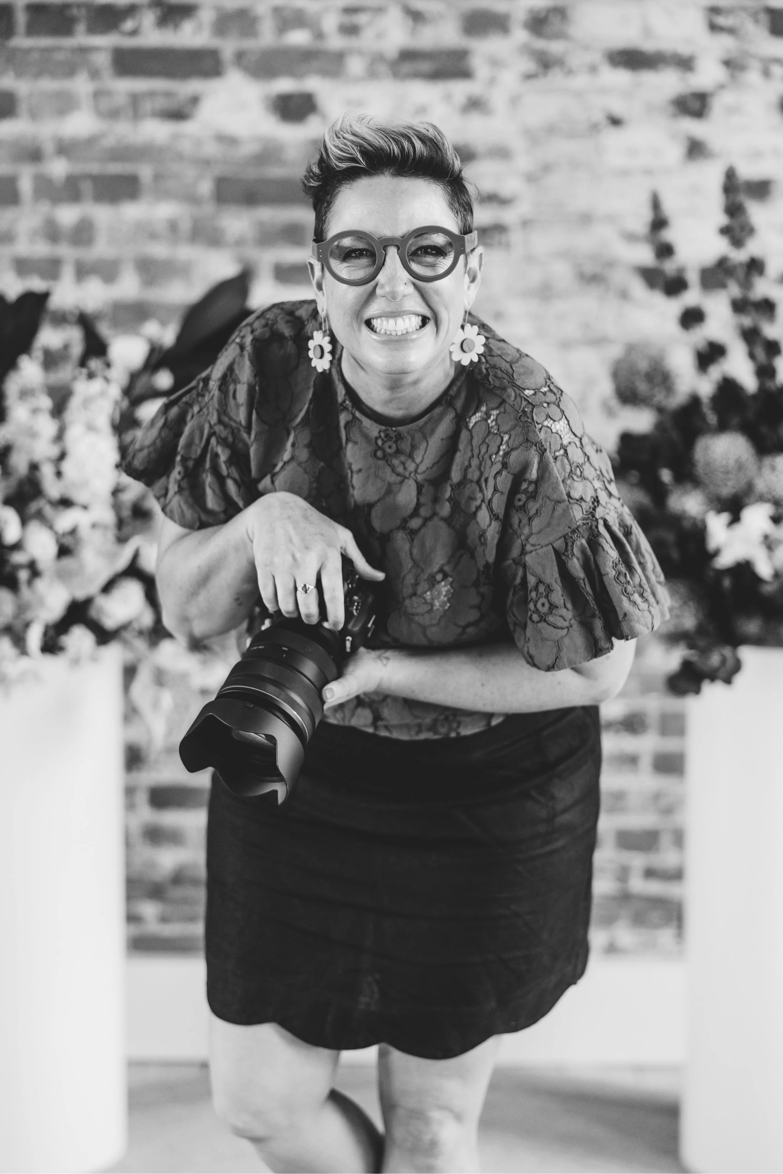 Smiling woman with shoulder-length black hair wearing a blue top, standing outdoors with green foliage in the background.