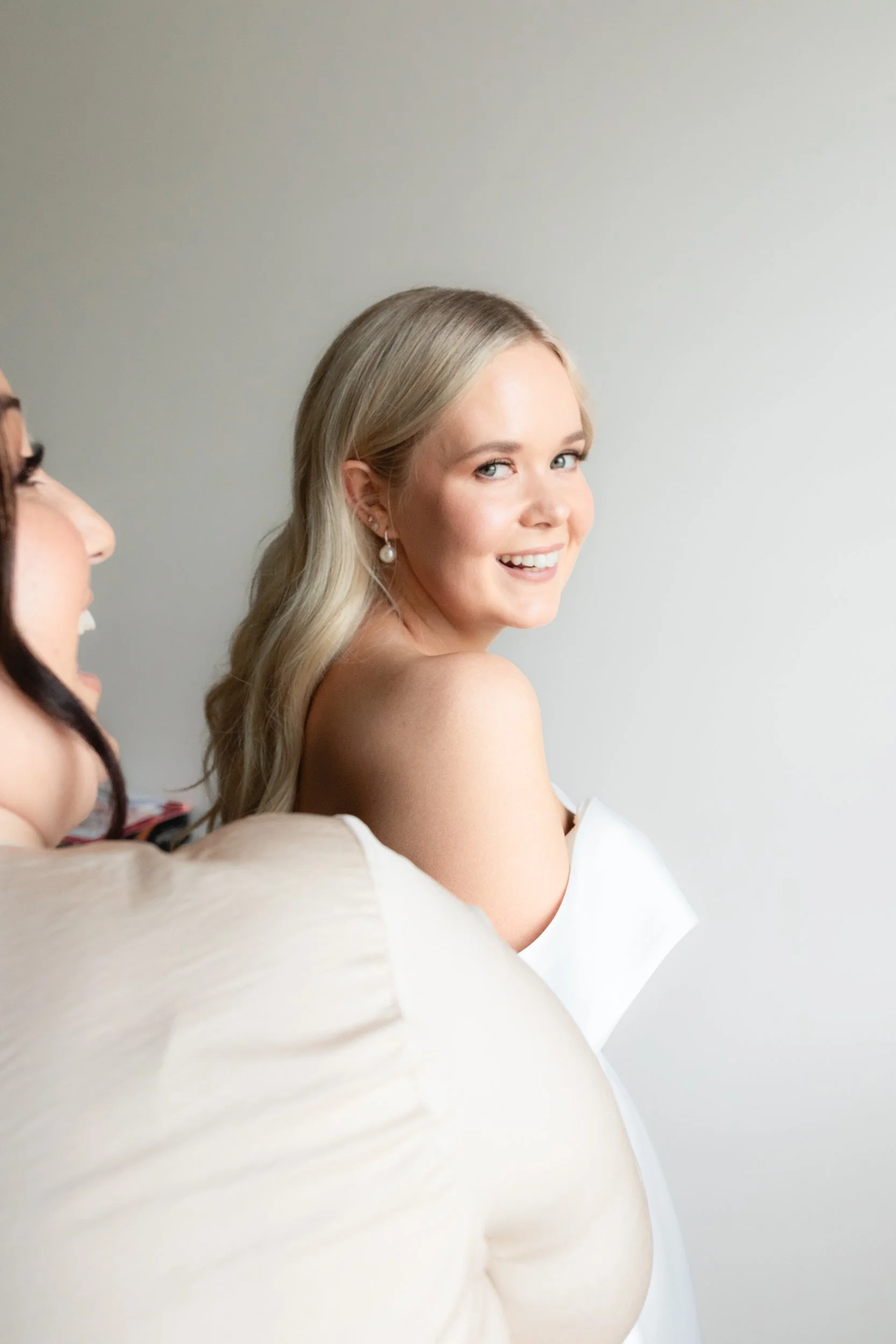 A young blonde woman smiling and looking over her shoulder, wearing pearl earrings and a white off-shoulder dress.