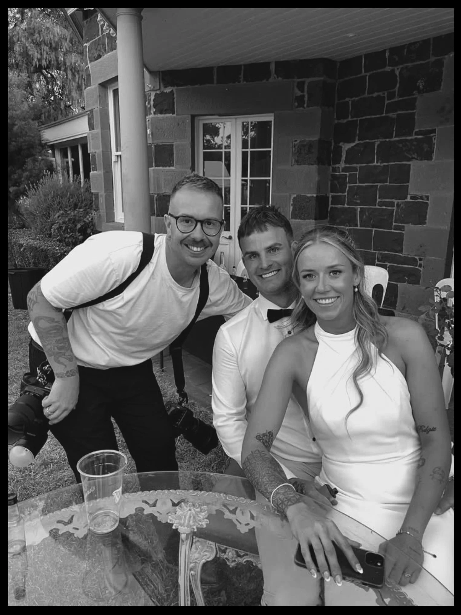 Tal with bride groom, at an outdoor gathering; the woman has long hair, tattoos, and a white dress, standing next to a man in a tuxedo and bow tie, while another man with glasses, tattoos, and a camera hangs around his neck leans in from the side.