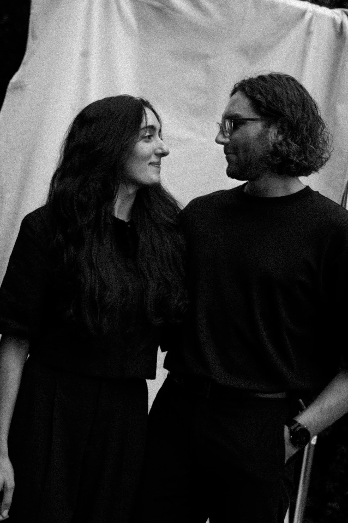 Black and white photo of All Souls Wedding. The woman has long, dark wavy hair. The man has shoulder-length curly hair, glasses, and a beard. They are in front of a plain backdrop.