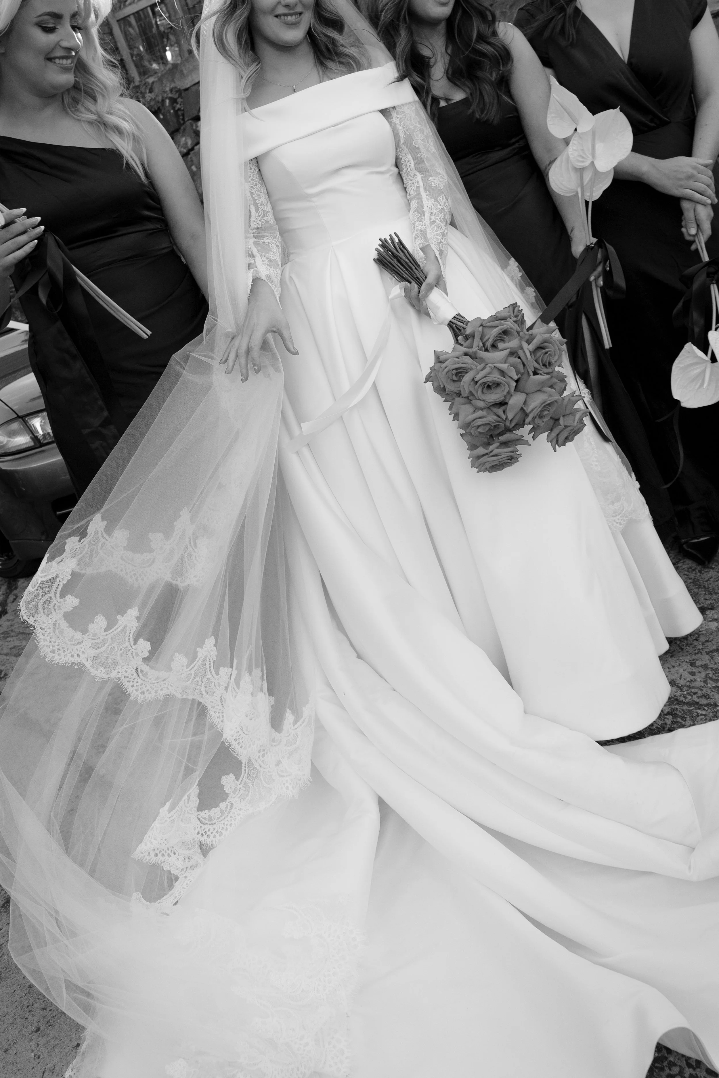 Black and white photo of a bride and her bridesmaids at a wedding. The bride is holding a bouquet of roses, wearing a wedding dress with lace sleeves and a veil, standing amid her bridesmaids who are dressed in dark dresses and holding white anthuriu