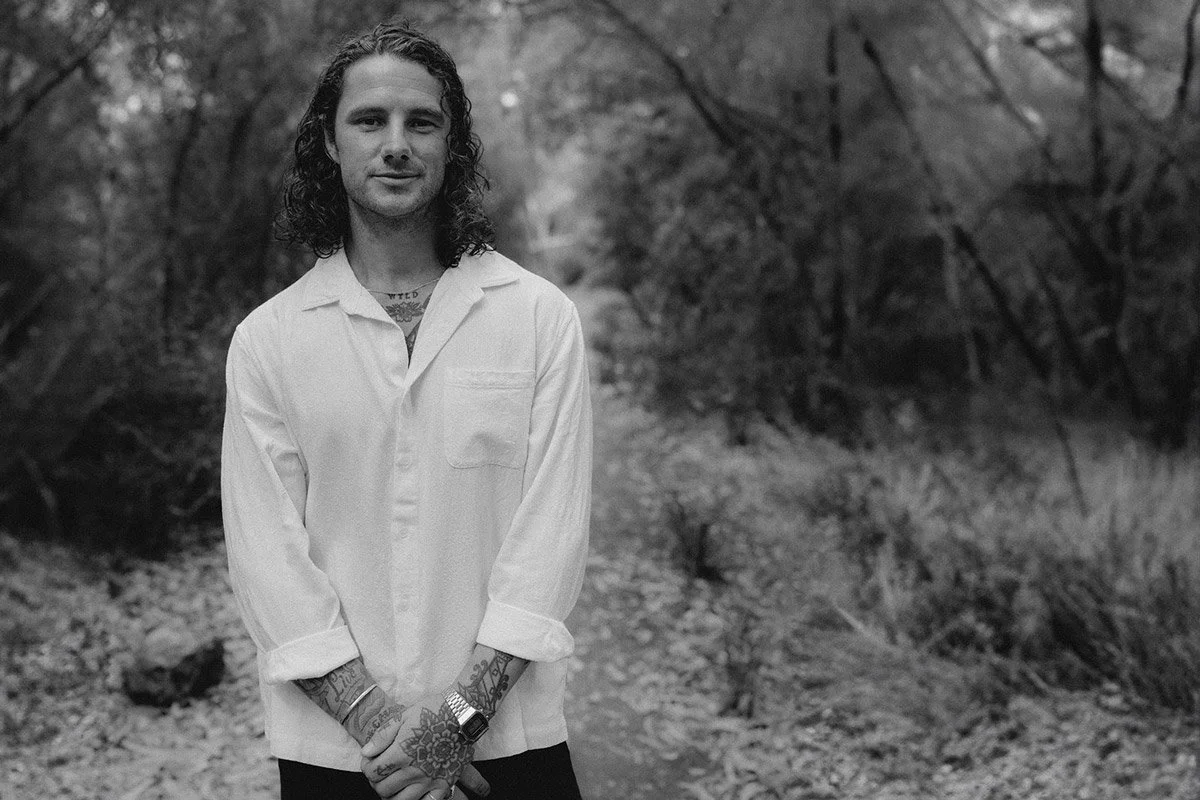 A man with long curly hair standing outdoors on a dirt path surrounded by trees, wearing a white button-up shirt with rolled-up sleeves, tattoos on his hands and neck, and a wristwatch.