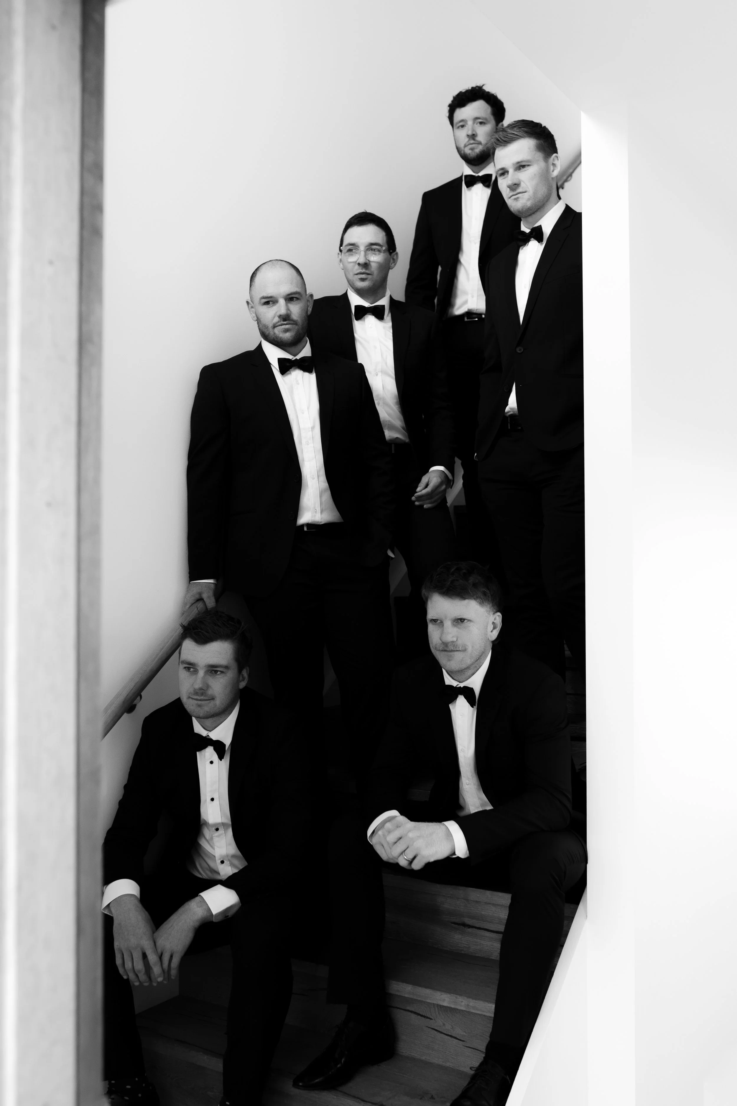Group of five men dressed in tuxedos, standing and sitting on a staircase, looking in different directions, in a black and white photo.