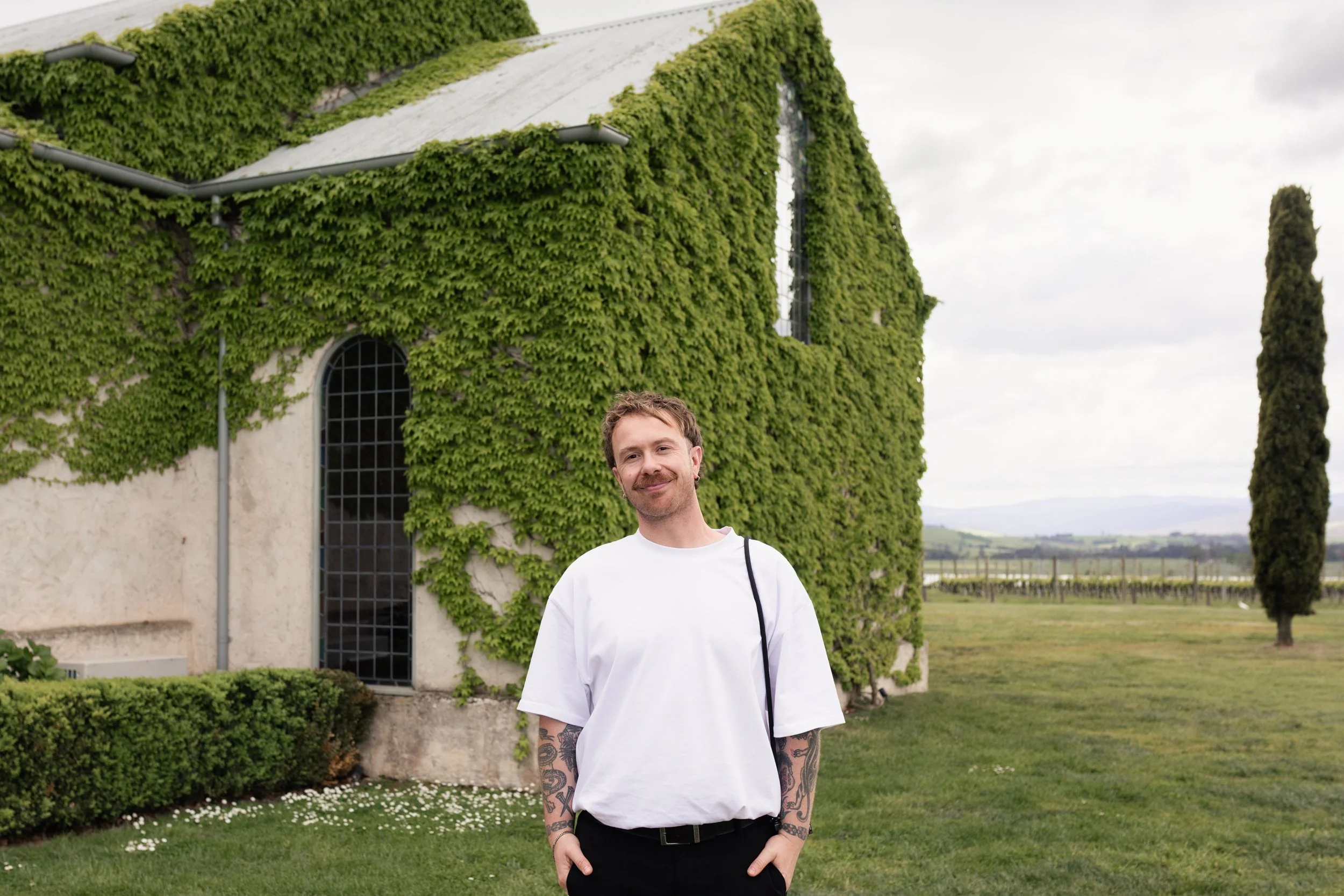 Tal Lemmens Wedding Photographer with tattoos on his arms, wearing a white T-shirt and black pants, standing outdoors in front of a building covered in green ivy, with a field and trees in the background.