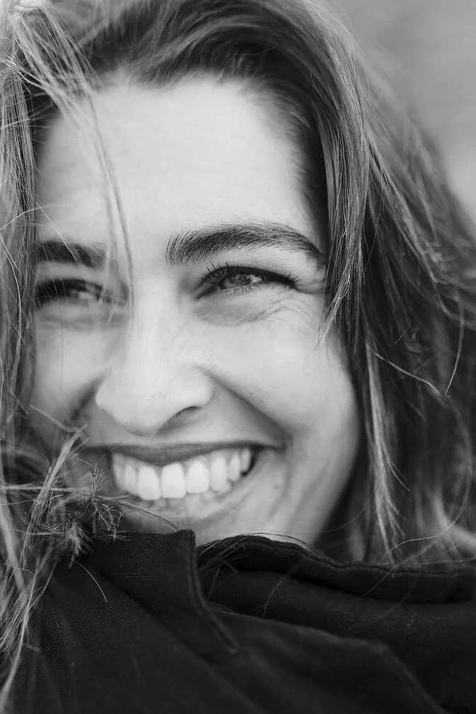 A man with curly dark hair, a beard, glasses, and a nose ring, looking slightly to the side with a neutral expression, inside a room with a blurred background.