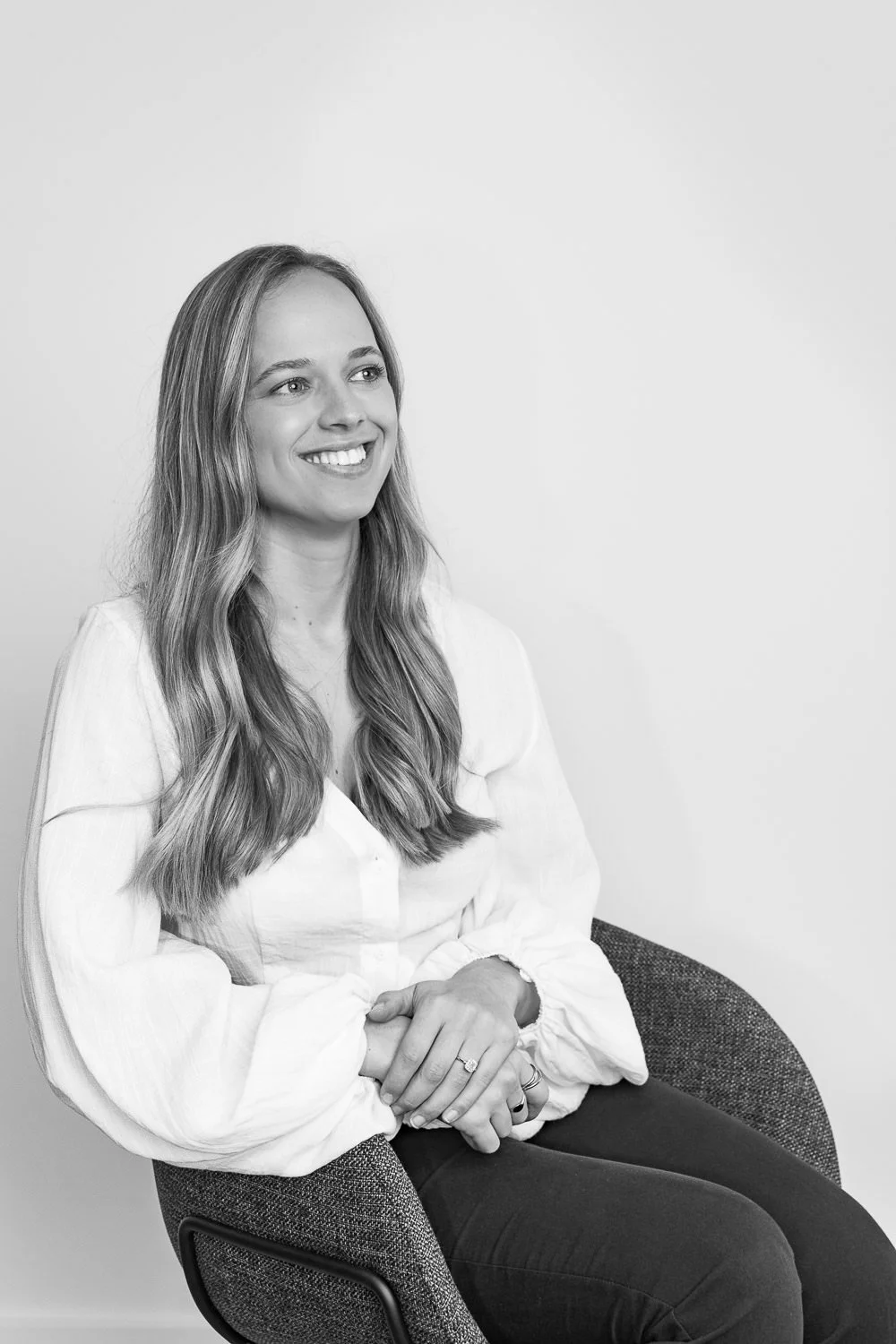 A black-and-white photo of a young woman with long, wavy hair smiling, wearing a light-colored blouse, sitting on a modern chair against a plain wall.