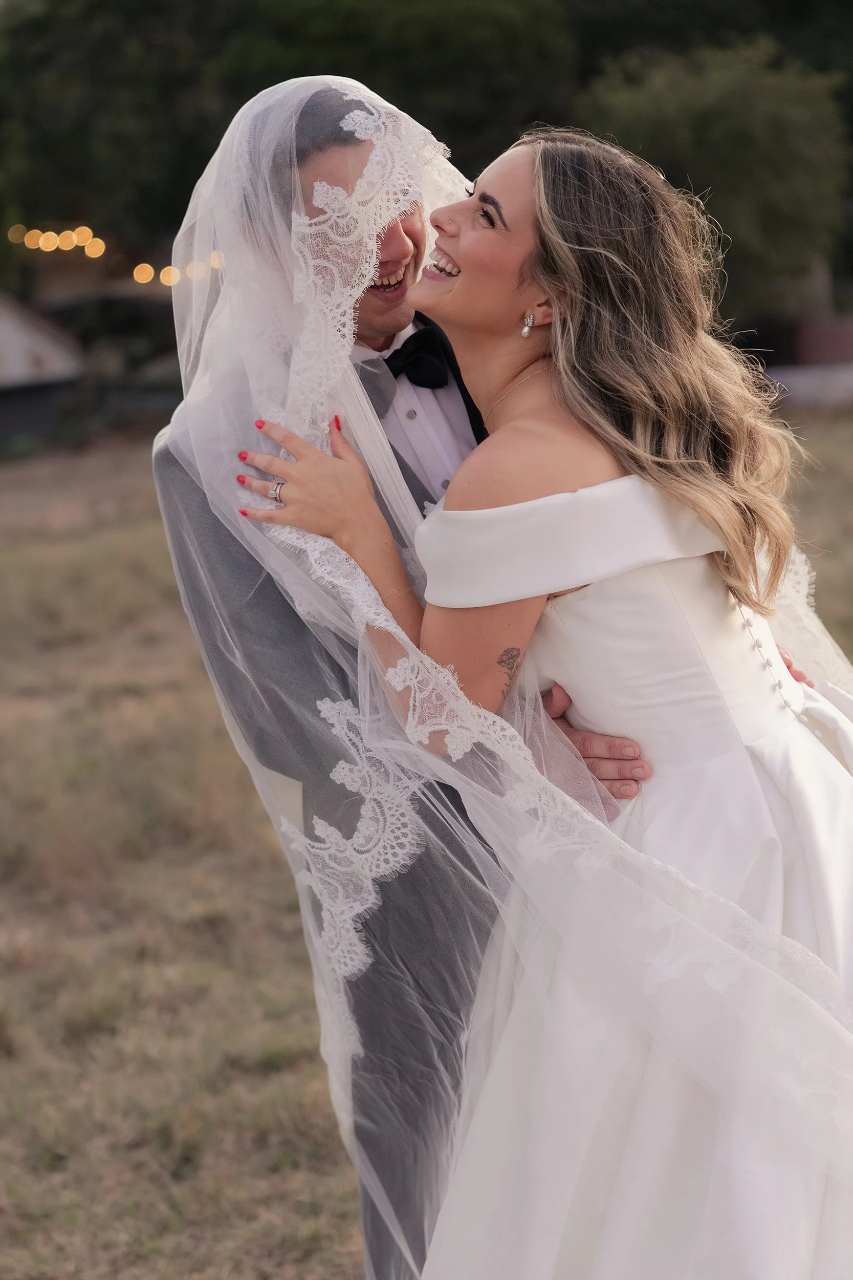 A bride and groom are smiling and embracing outdoors, with the bride wearing an off-shoulder white dress and pearl earrings, and the groom in a tuxedo with a black bow tie, the bride's hand showing an engagement ring and red nail polish.