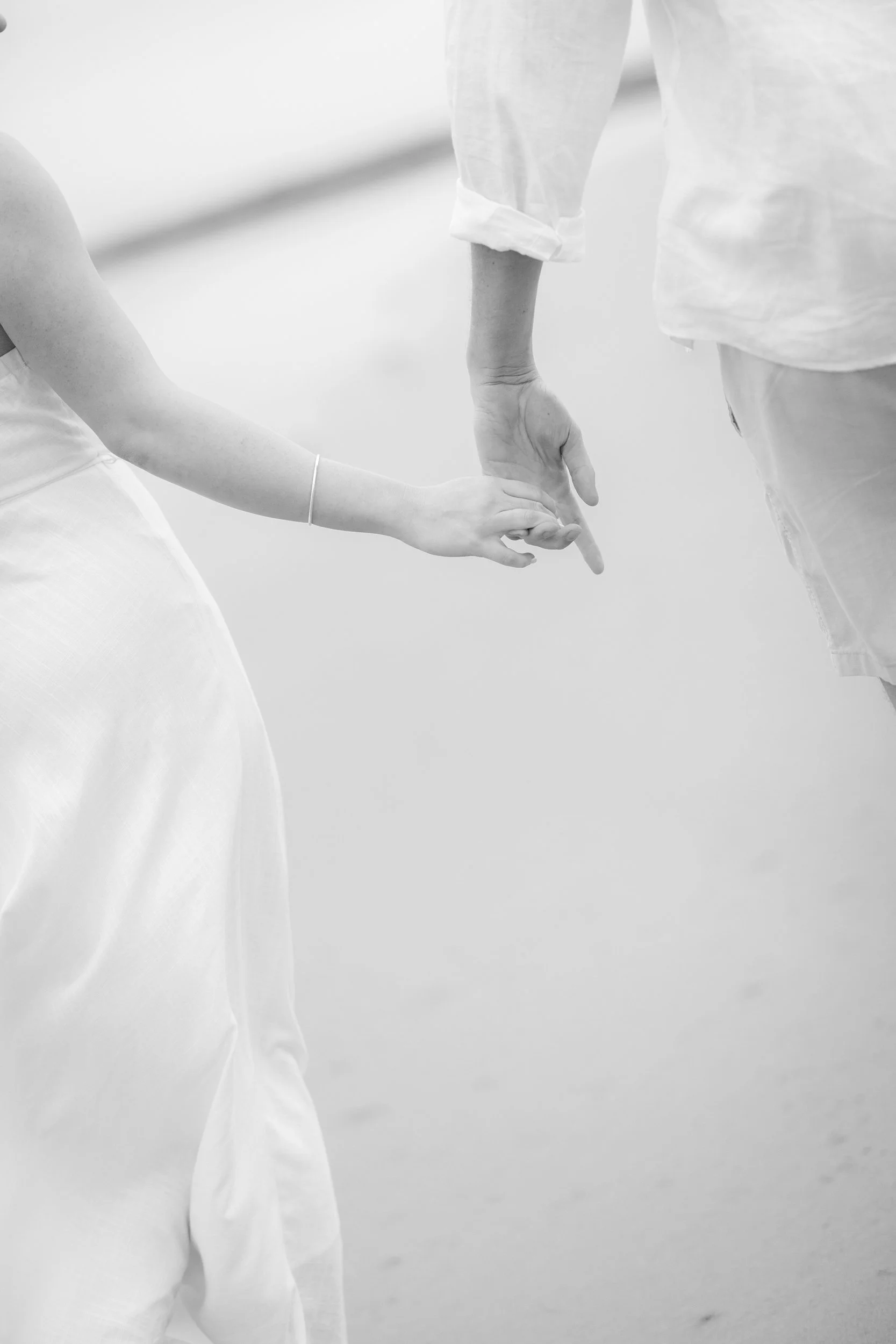Close-up of two engaged partners holding hands, one with an outstretched arm and the other with a relaxed hand, both wearing light-colored costumes.