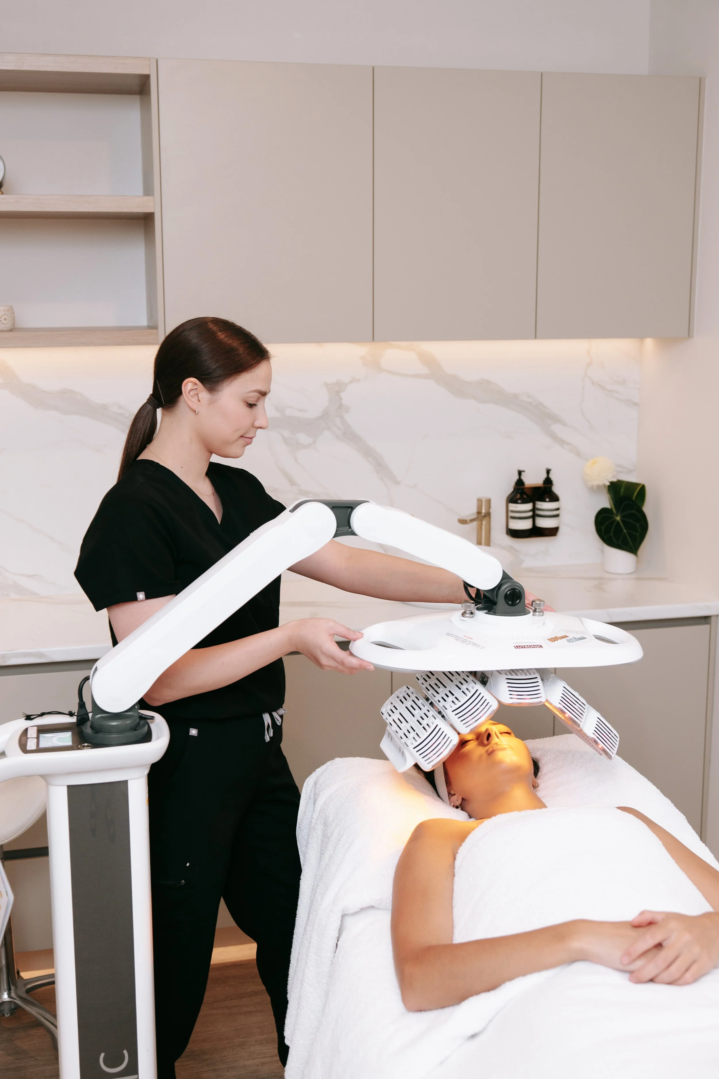A woman relaxing during a facial treatment in a spa or skincare clinic, with a facial steamer positioned above her face, while a professional technician waits on the side.