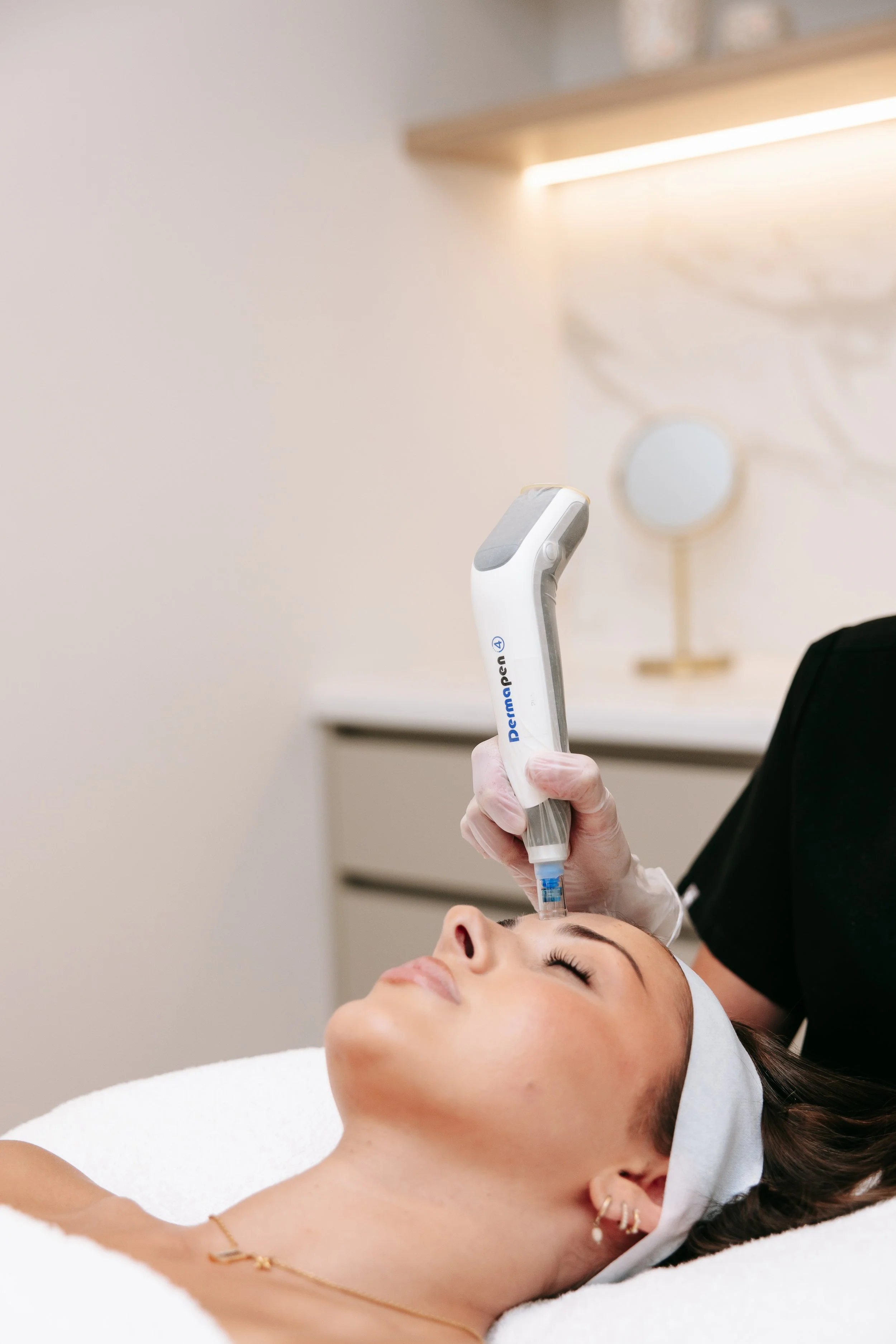 A woman lying on a treatment bed with her eyes closed, receiving a facial treatment from a professional using a handheld device, in a spa or clinic setting.