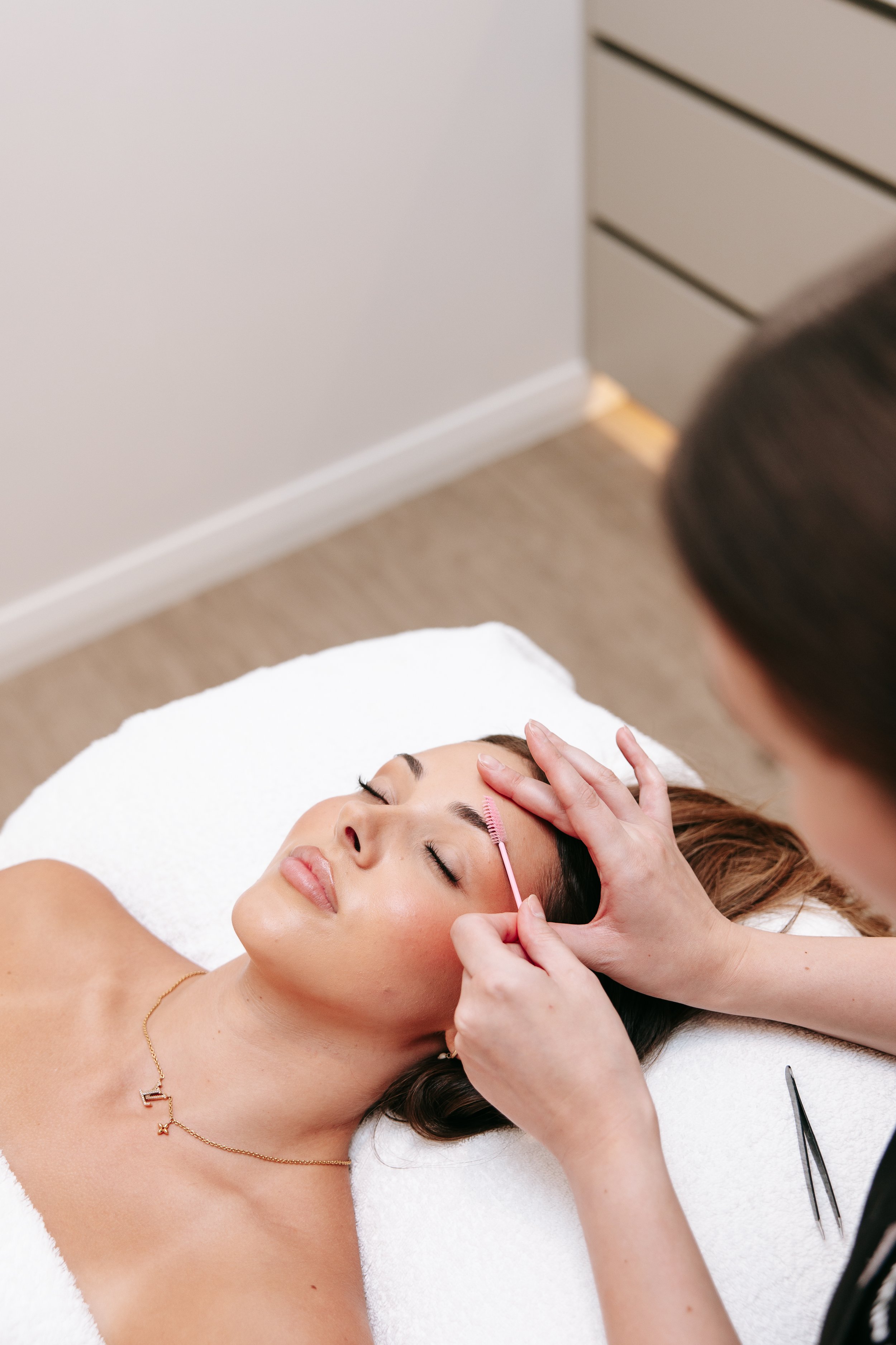 A woman is lying down with her eyes closed, receiving a facial treatment from an esthetician. The esthetician is gently touching her forehead and cheeks during the procedure.