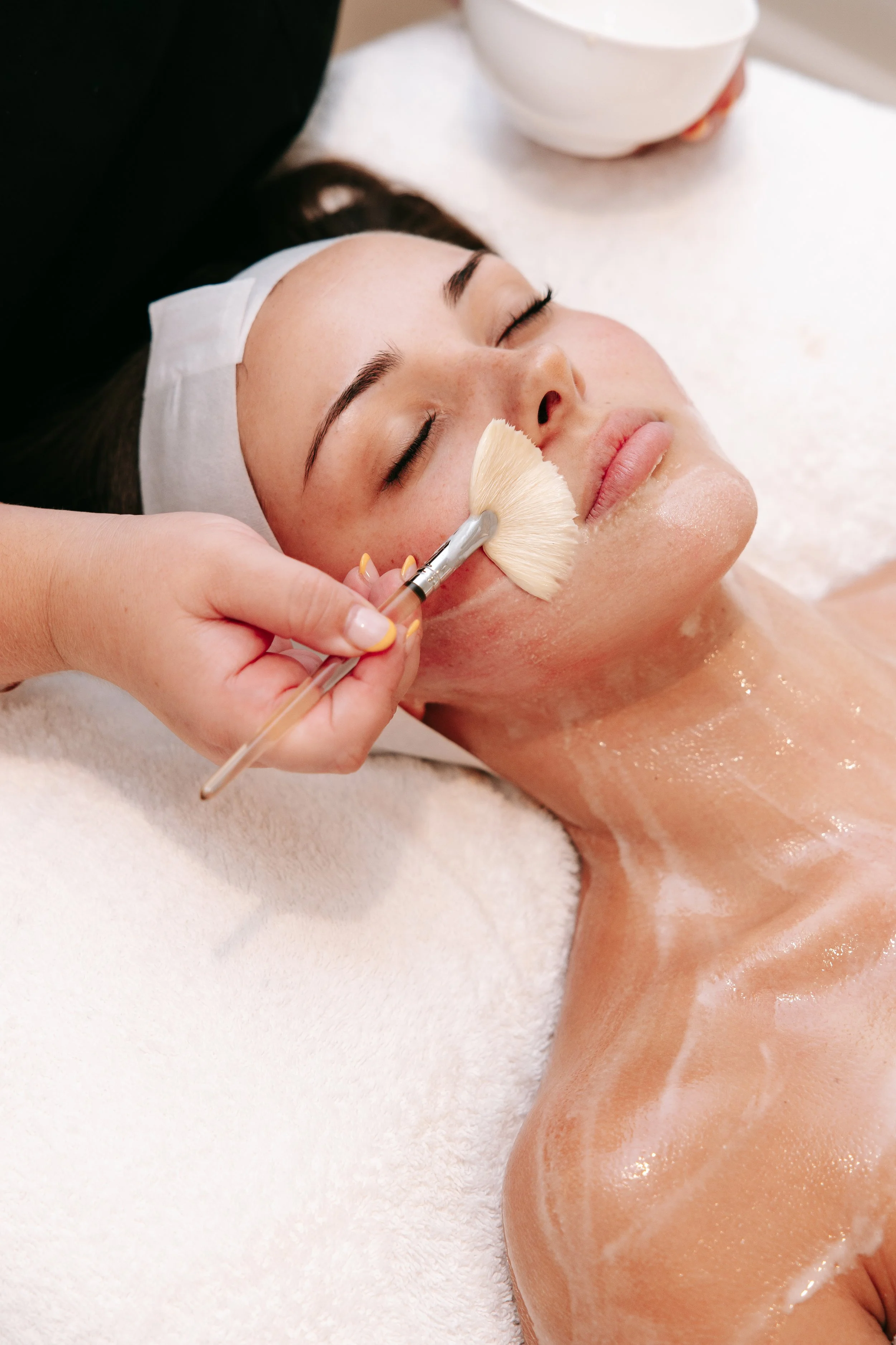 A woman receiving a facial treatment, with her eyes closed, lying on a treatment bed with a white towel, as an aesthetician applies a facial mask or scrub using a fan brush. There are white bowls nearby.