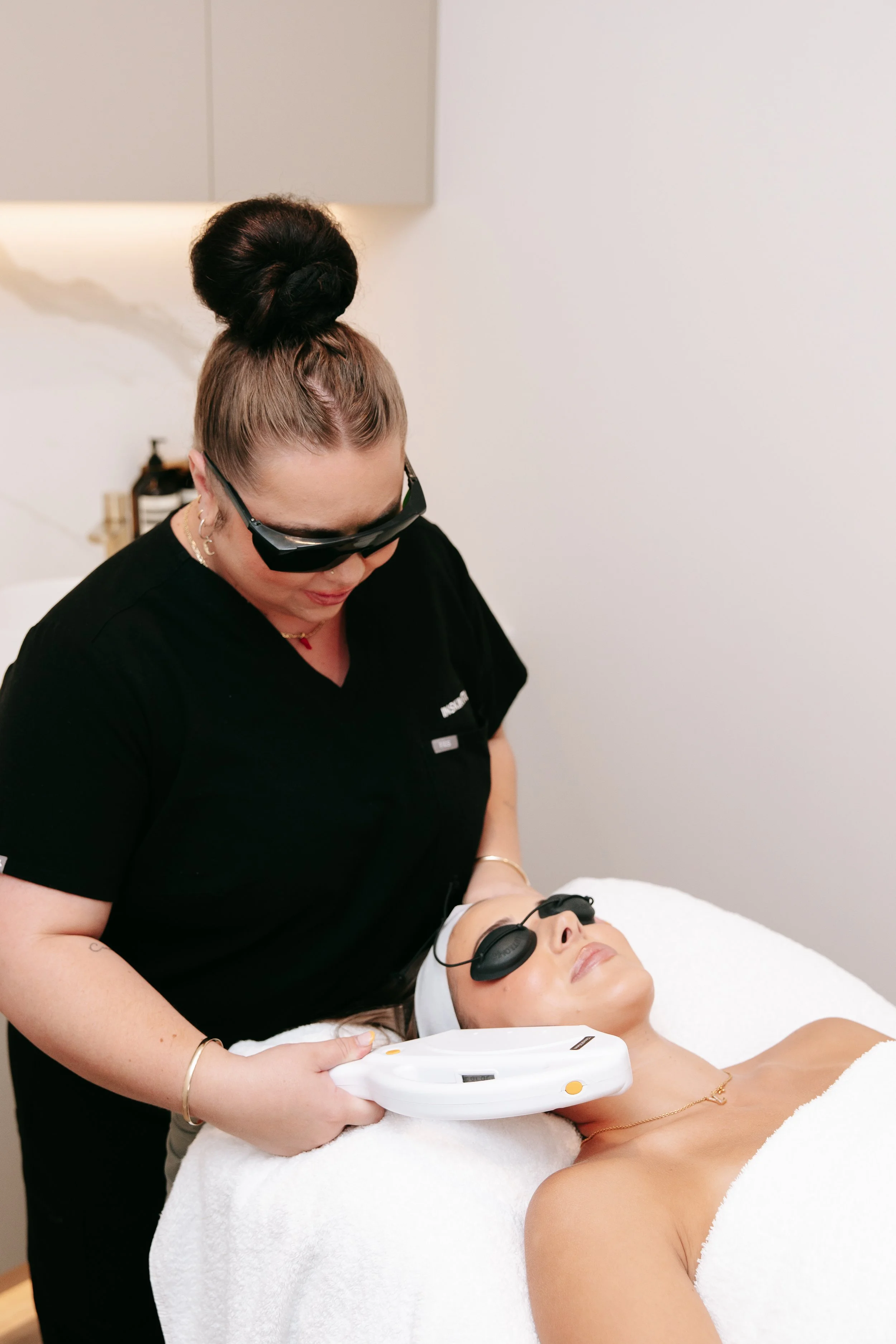 A woman receiving a facial treatment from a professional in a spa or clinic setting. The practitioner is wearing glasses and a black uniform, and the client is lying down with protective eye covers, relaxing with closed eyes.