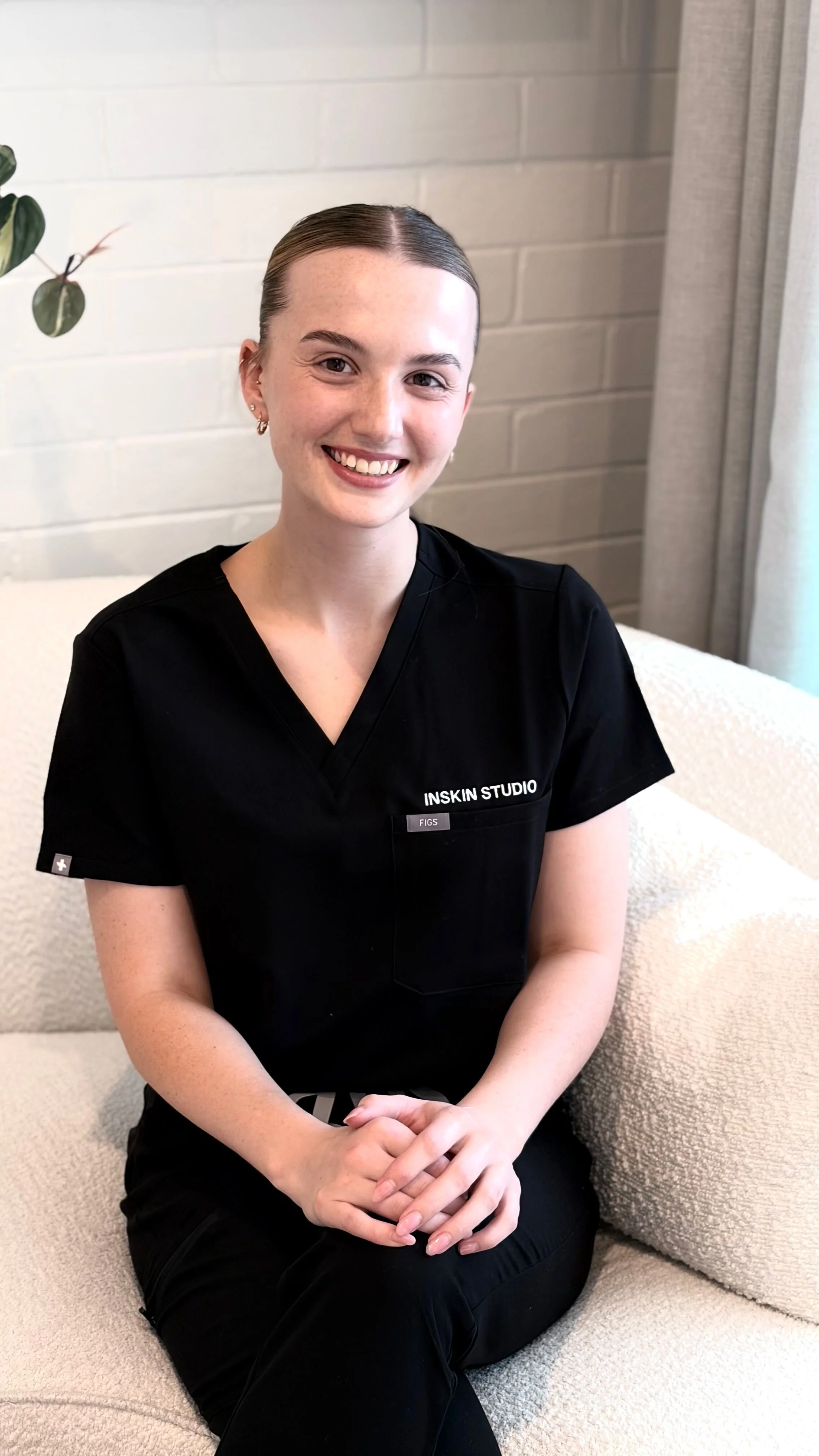 A young woman with short brown hair sitting on a beige couch, smiling, wearing a black uniform with 'INSKIN STUDIO' embroidered on it, in a room with a brick wall and a plant in the background.