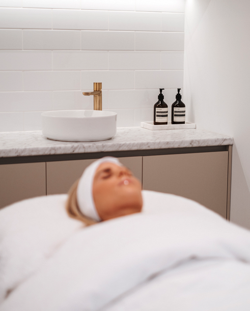 A woman lying in a bed with a white headband in a spa or treatment room, with a sink and soap dispensers on the countertop behind her.