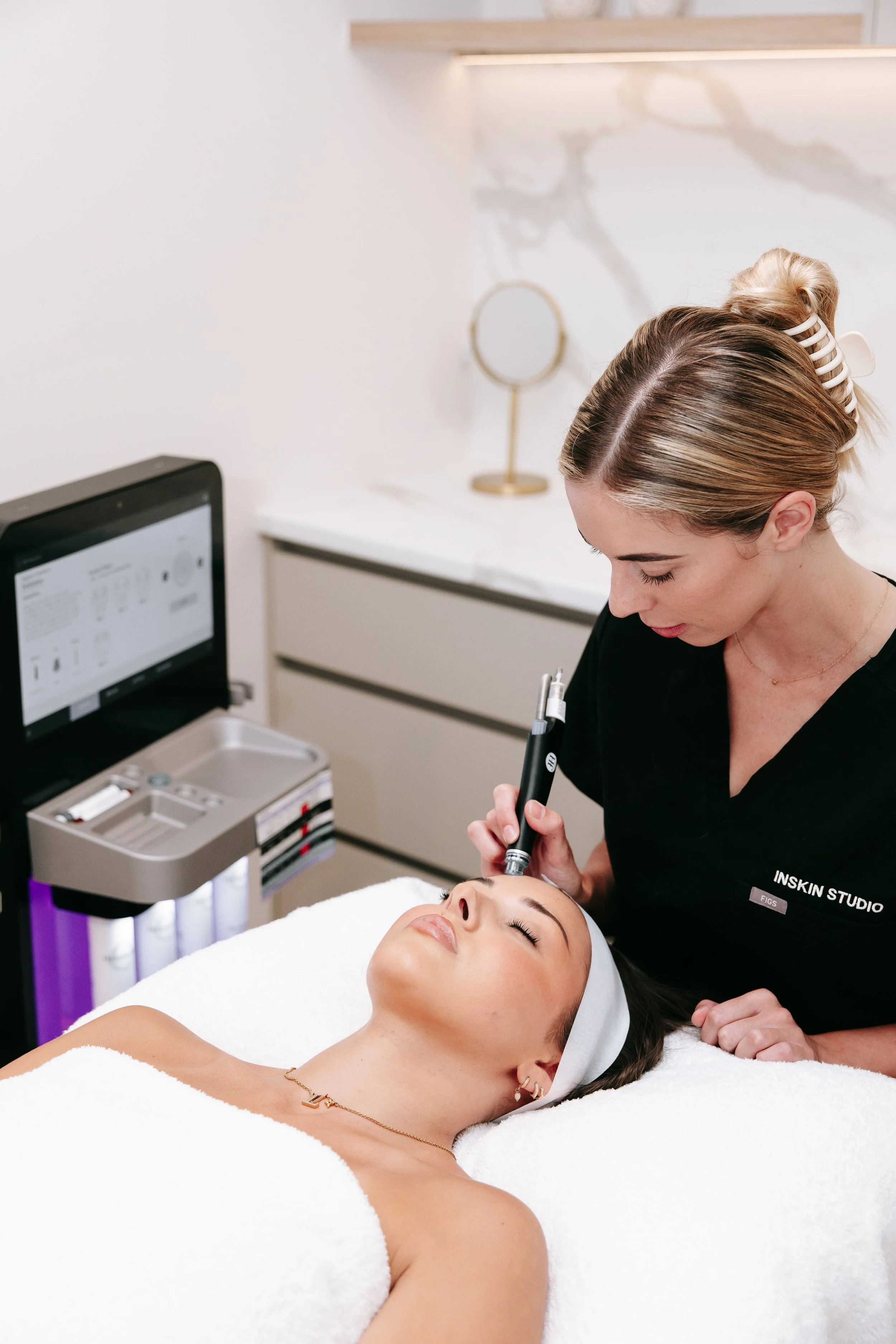A woman is lying down with her eyes closed, receiving a facial treatment from a skincare professional in a spa or clinic setting. The skincare professional is holding a device close to the woman's face, and there are various skincare products or equipment nearby.