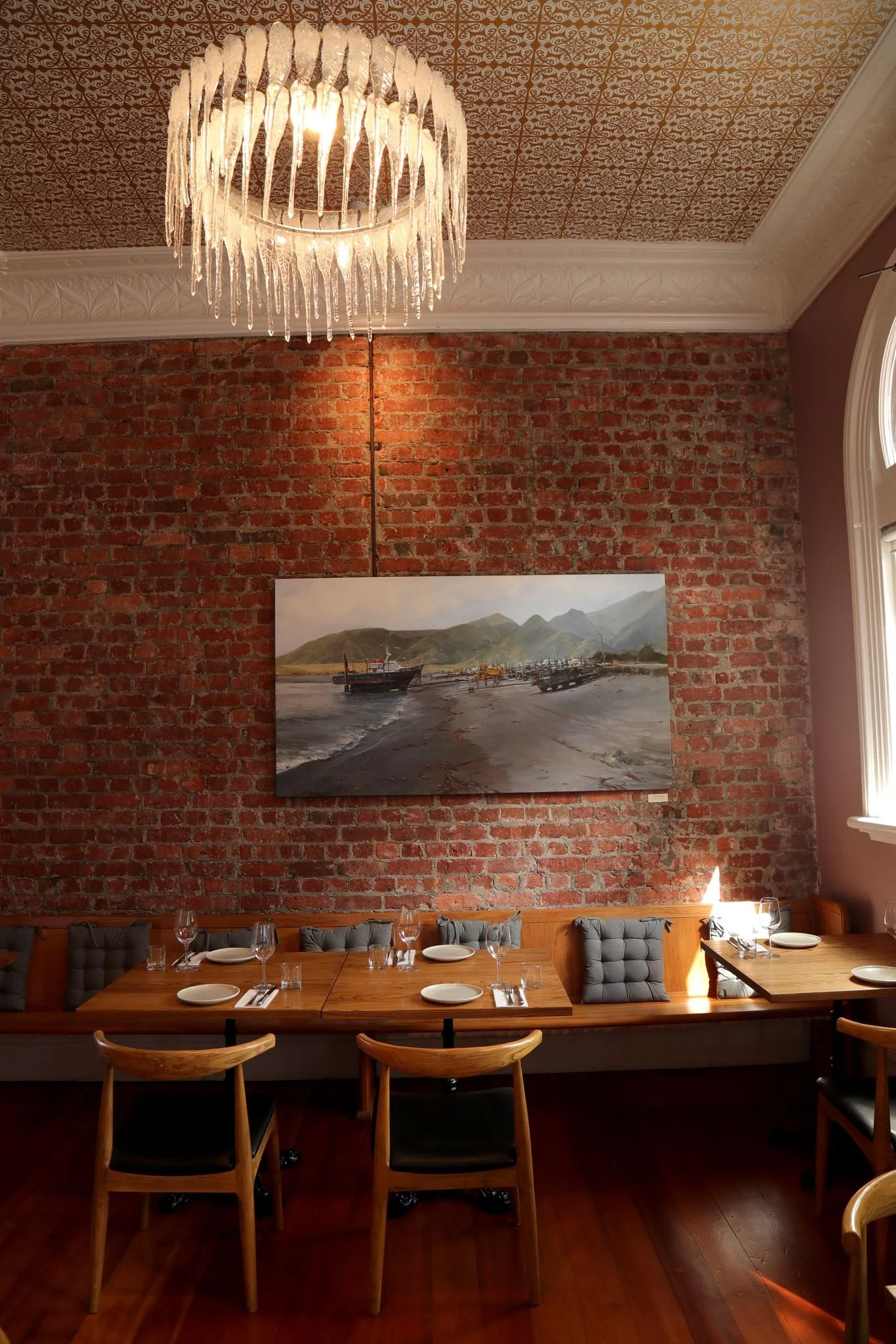 A cozy restaurant interior with an exposed brick wall, a landscape painting of boats on water and mountains, wooden tables set with plates and glasses, a chandelier with hanging glass pieces, and a large window letting in natural light.