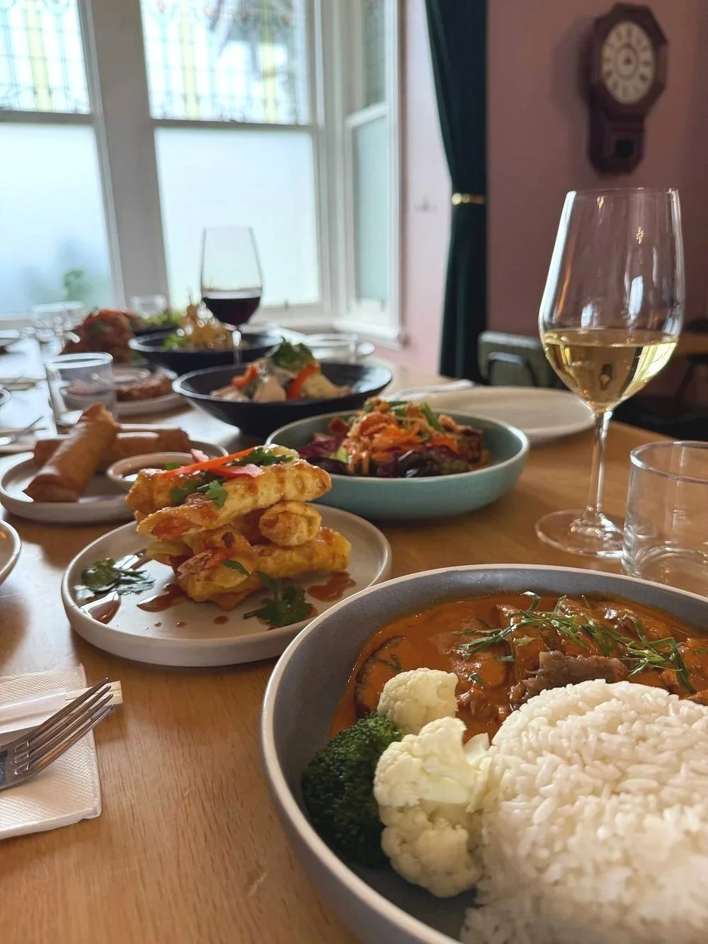 A table set with a variety of dishes including a bowl of rice and curry with broccoli, a plate of fried appetizers garnished with herbs, various salads, a glass of white wine, and a glass of red wine, with an open window and a wall clock in the background.