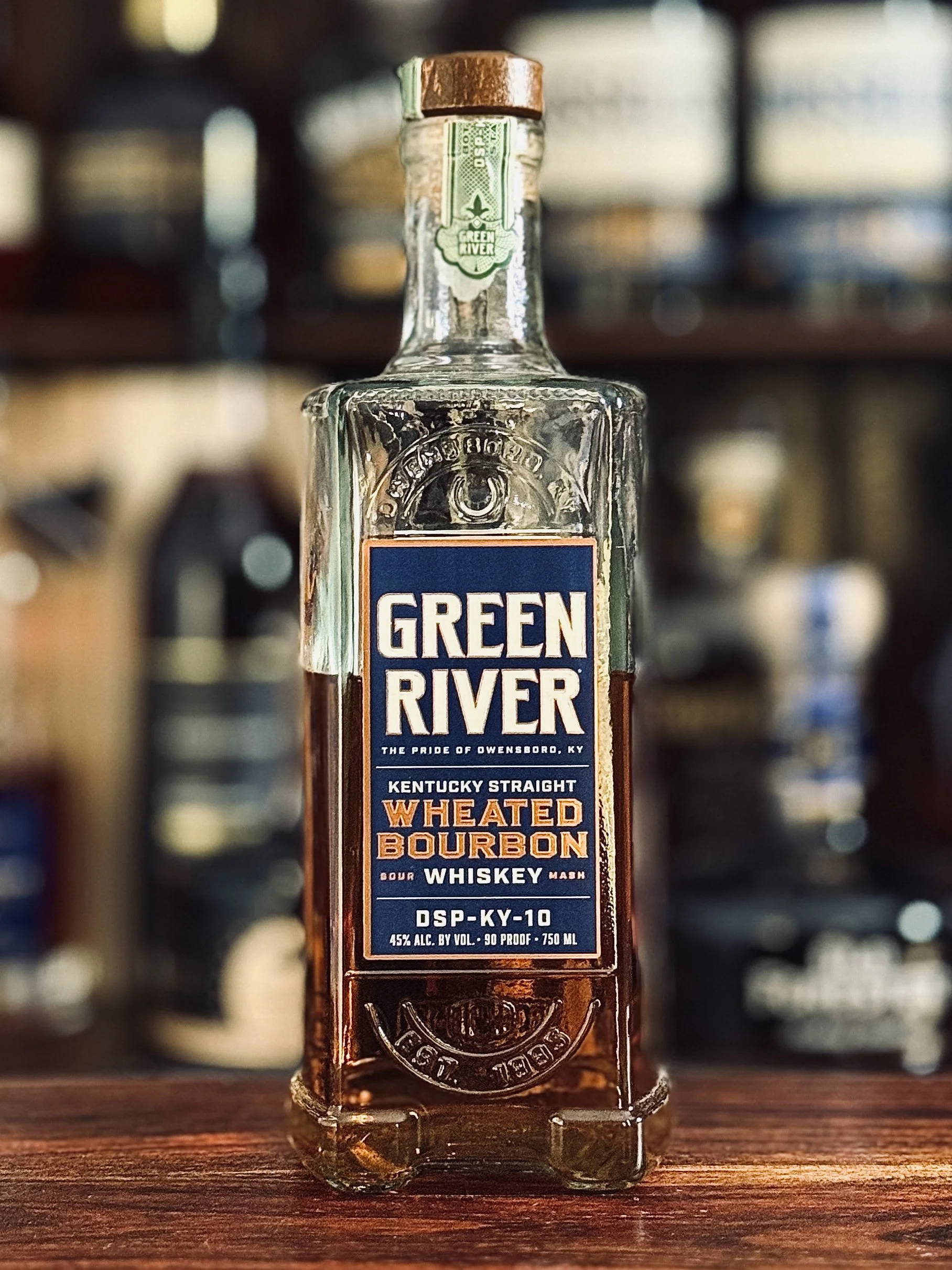 Green River Kentucky Straight Wheated Bourbon bottle displayed on a wooden bar top, with warm lighting and a blurred whiskey collection behind it.