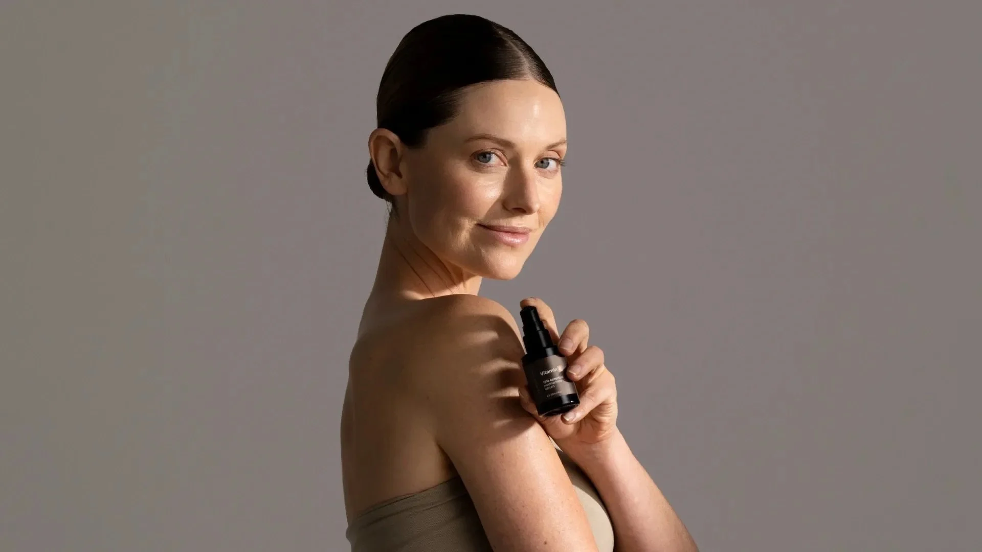 A woman with short dark hair holding a black skincare spray bottle on her shoulder, smiling softly against a neutral background. Dermiotic by Synergie Skin.