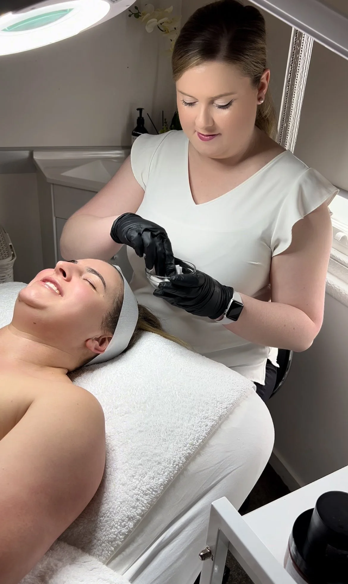A woman with a headband lying on a treatment bed, smiling with eyes closed, as a skincare specialist in black gloves applies a facial treatment using a small tool. The scene is in a spa or clinic room.
