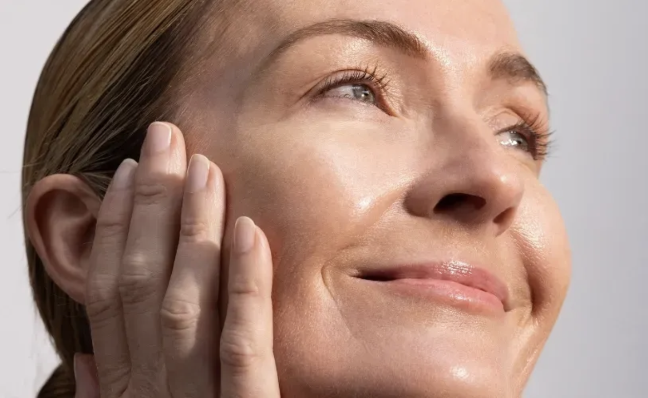 Close-up of a woman with smooth, glowing skin gently touching her face, smiling softly with eyes looking upward.